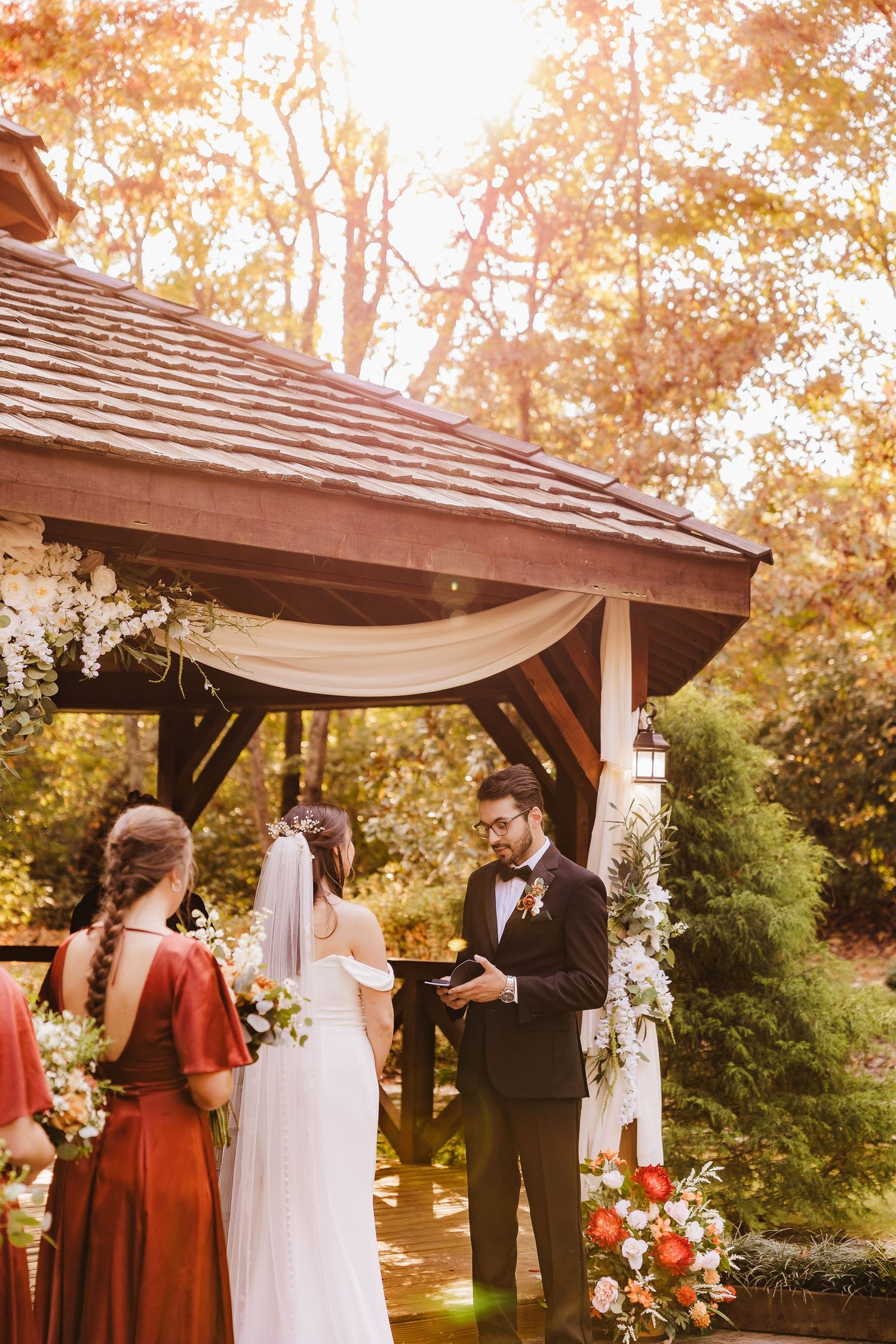 Wedding ceremony taking place under a wooden gazebo at sunset with autumn trees in the background.