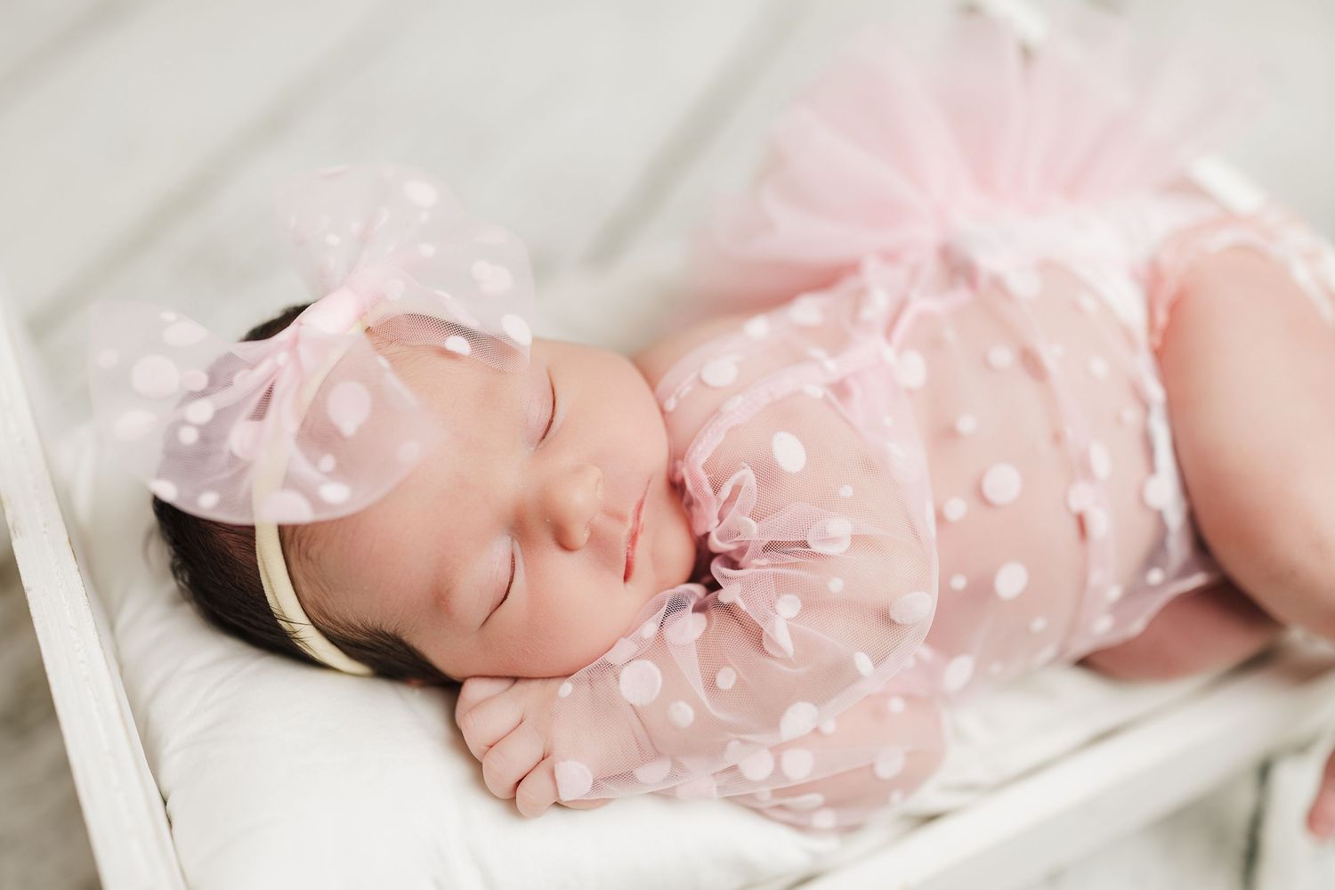 Sleeping baby wearing delicate pink lace dress with bow headband rests peacefully on white wooden surface.