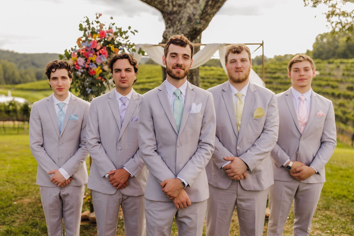 Five groomsmen in light gray suits with pastel ties pose outdoors at a vineyard wedding ceremony.