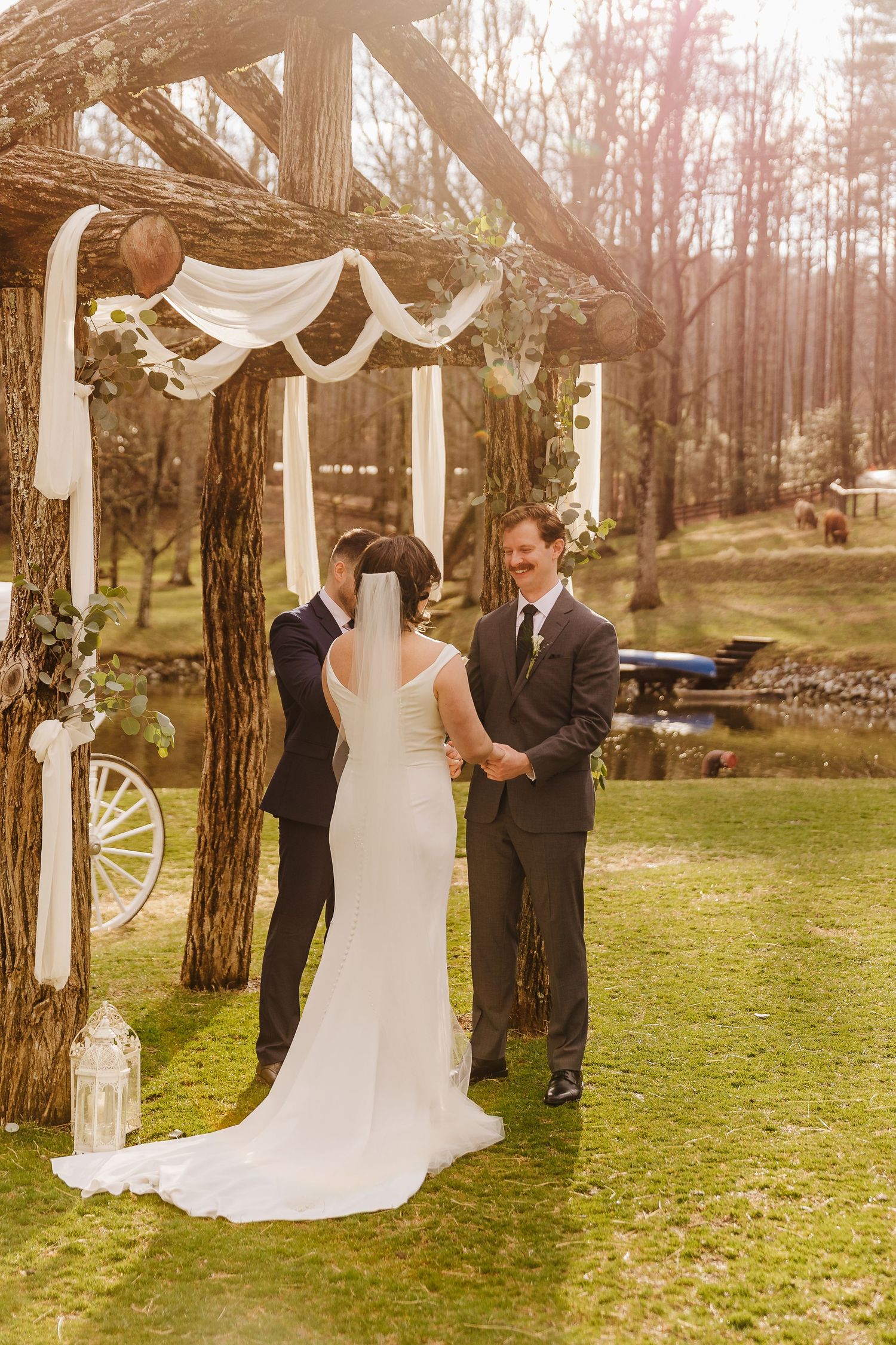 Bride and groom exchange vows outdoors under a rustic wooden arch draped with white fabric at a woodland wedding ceremony.