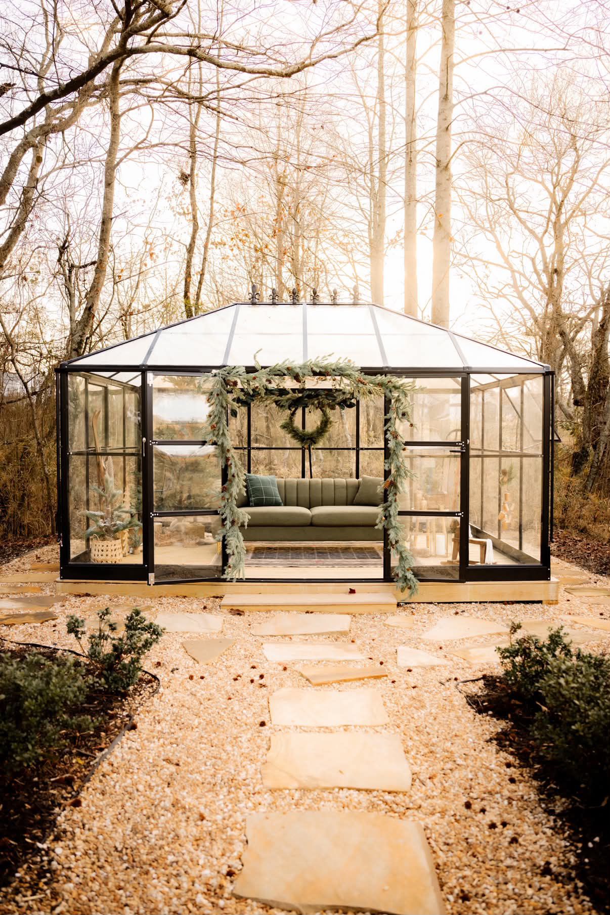 Glass greenhouse with green sofa, wreath, and garland decor surrounded by bare trees on a stone path.