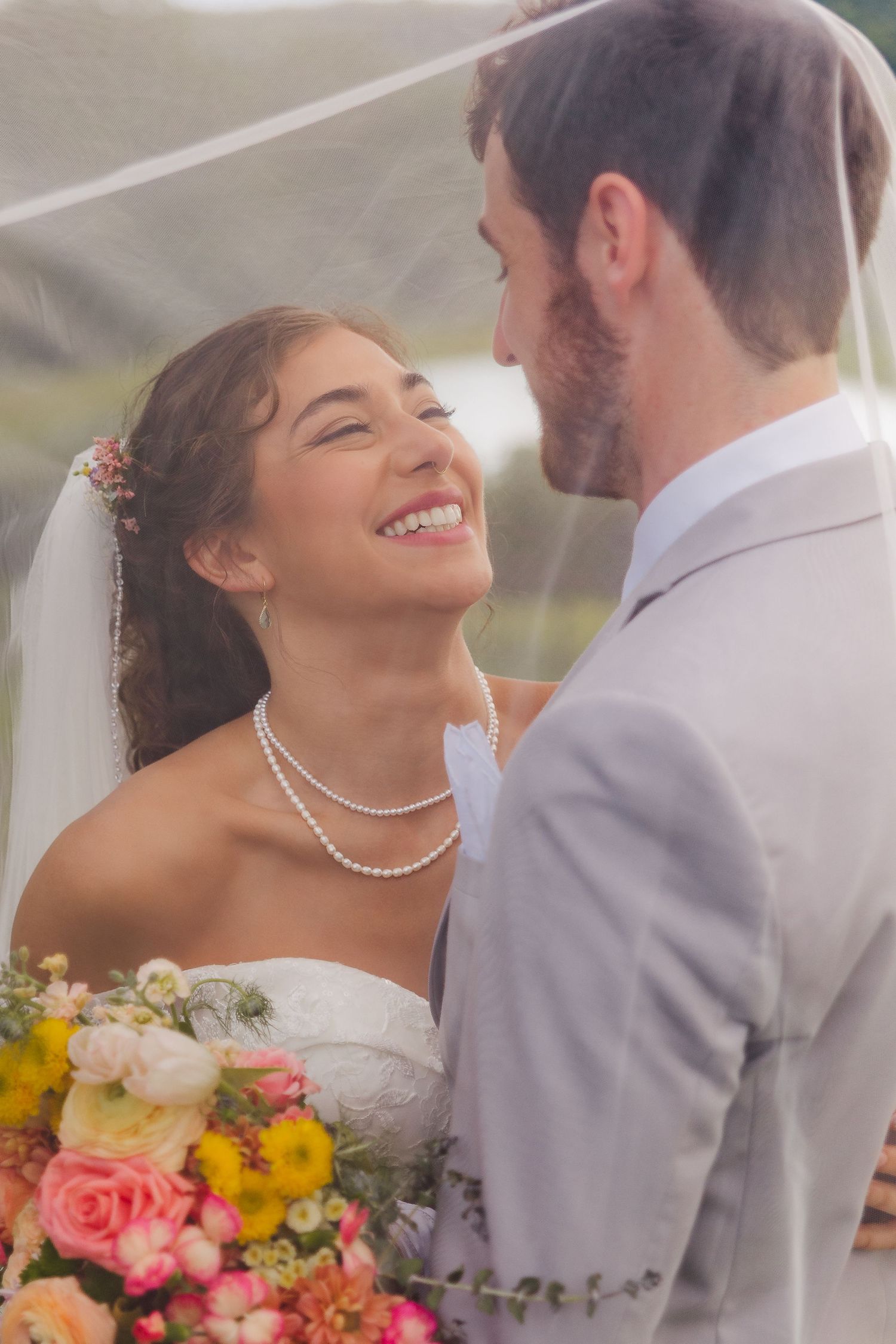 Smiling bride in white dress and pearl necklace holds colorful bouquet while gazing at groom in gray suit.
