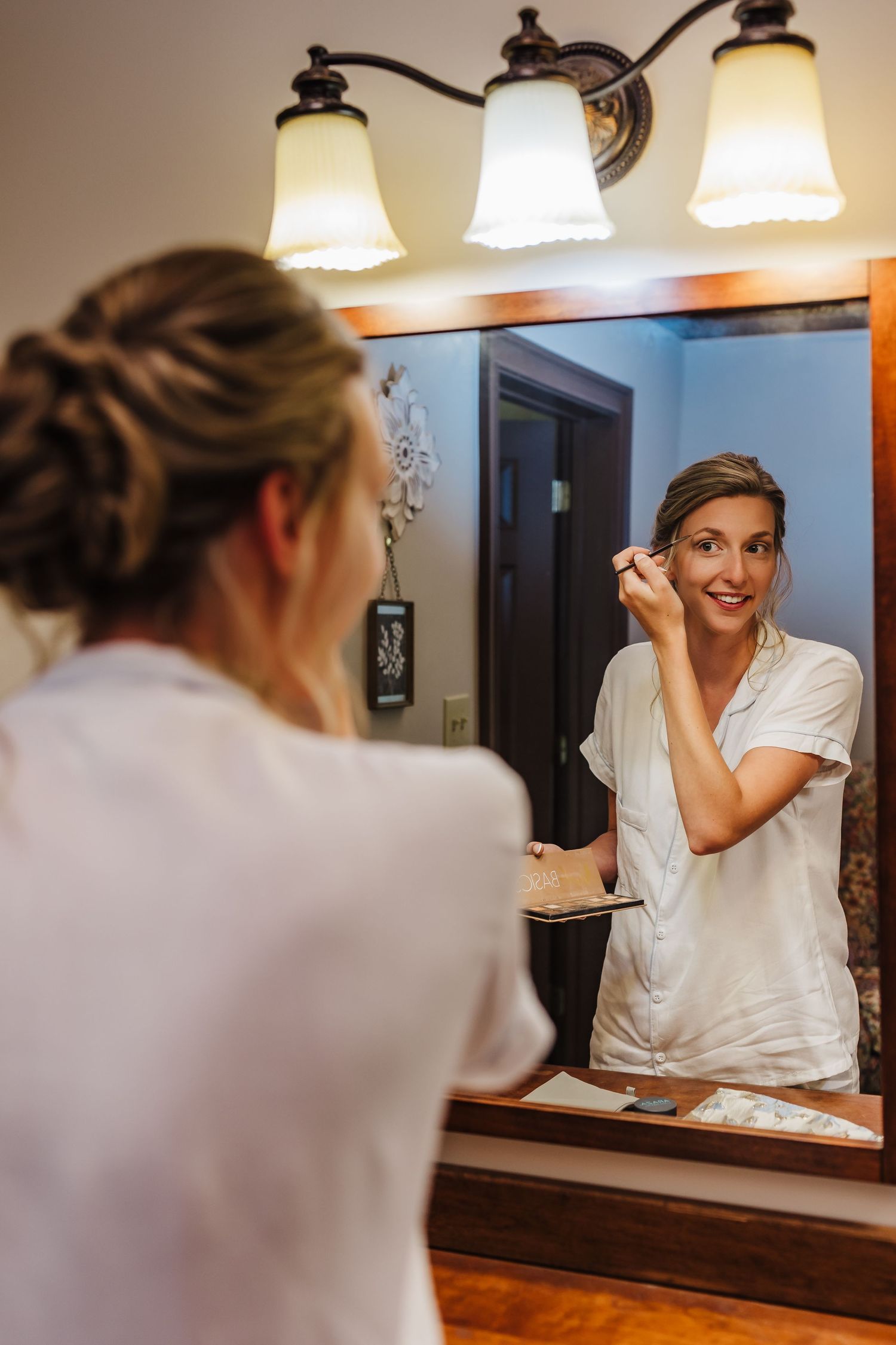 Bride getting ready with a mirror reflection in a bathroom at Walters Barn, featuring vintage-style lighting fixtures.