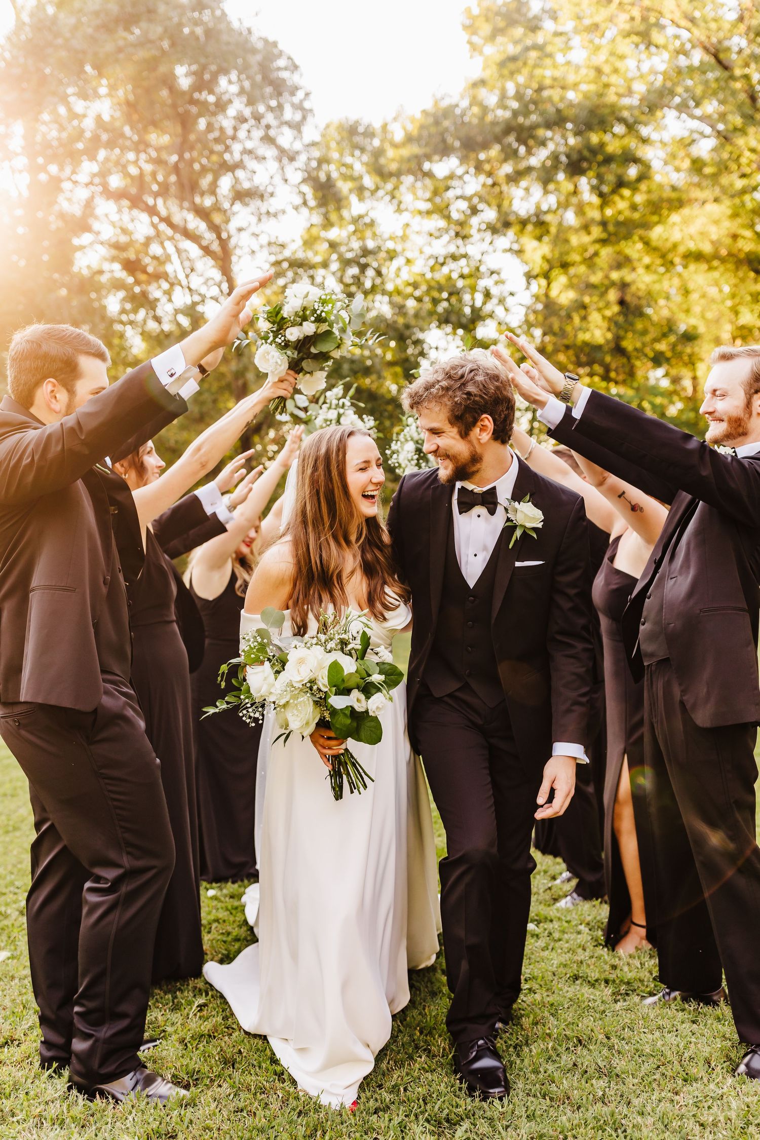 Wedding party in formal attire celebrating with raised arms outdoors at sunset.