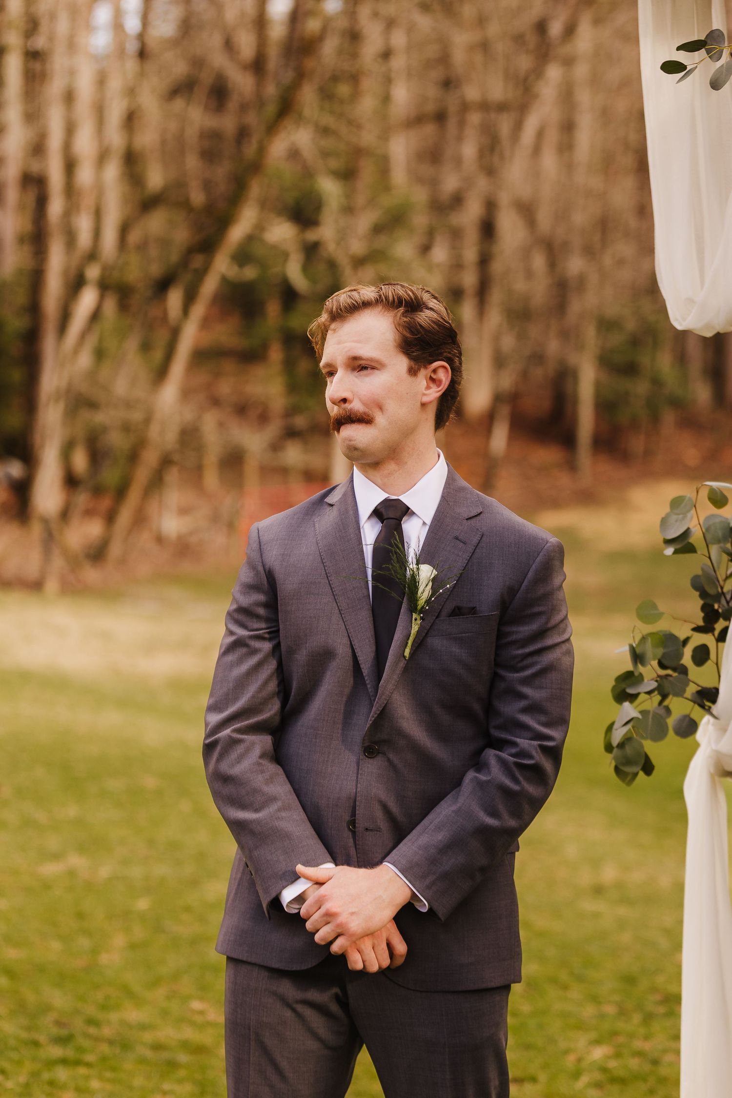 Groom in gray suit with boutonniere stands at outdoor wedding altar surrounded by trees.