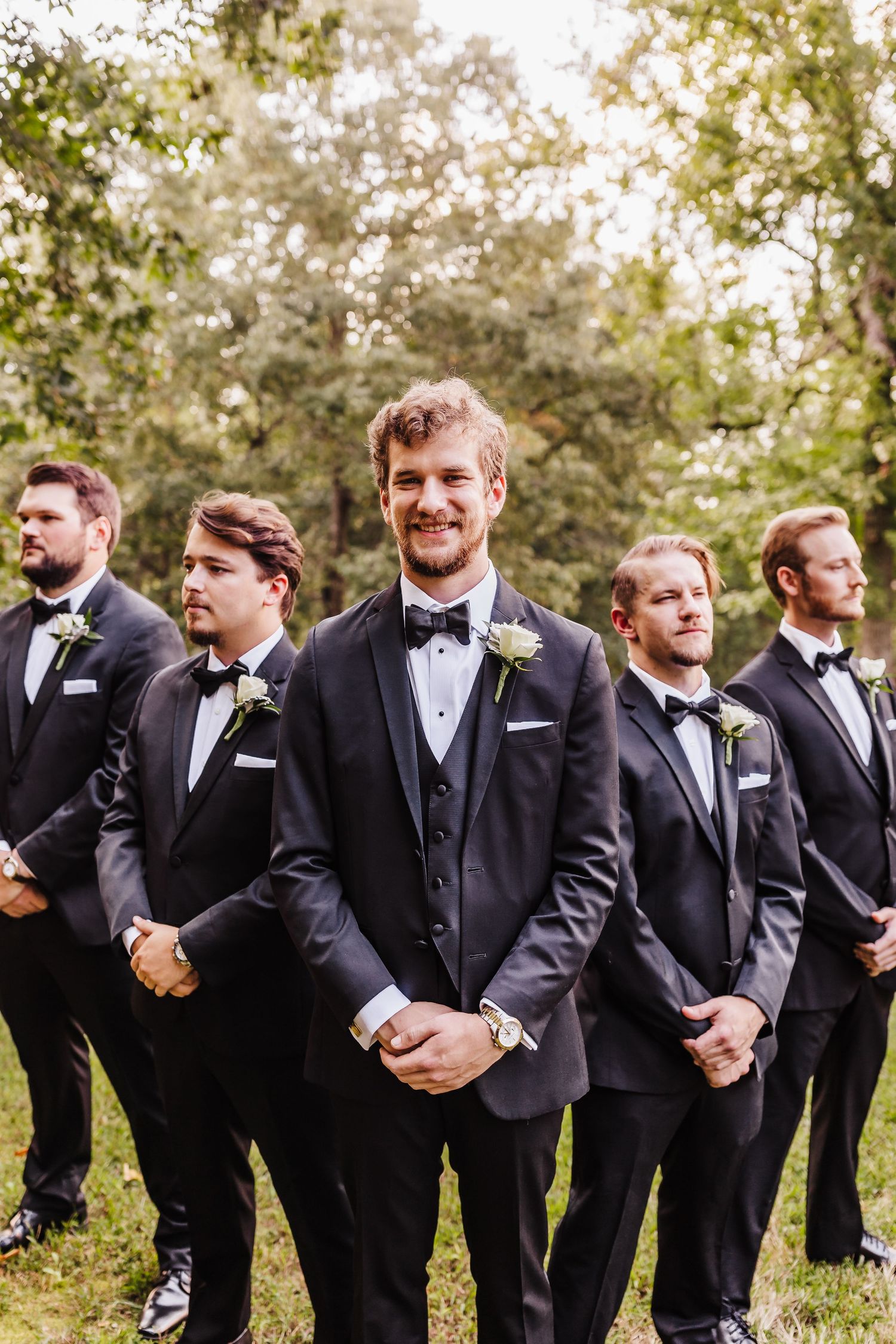 Groomsmen in black tuxedos posed outdoors in natural woodland setting.