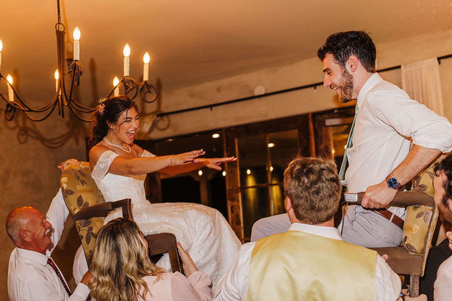 Bride and groom lifted on chairs during traditional Hora dance at elegant candlelit wedding reception.