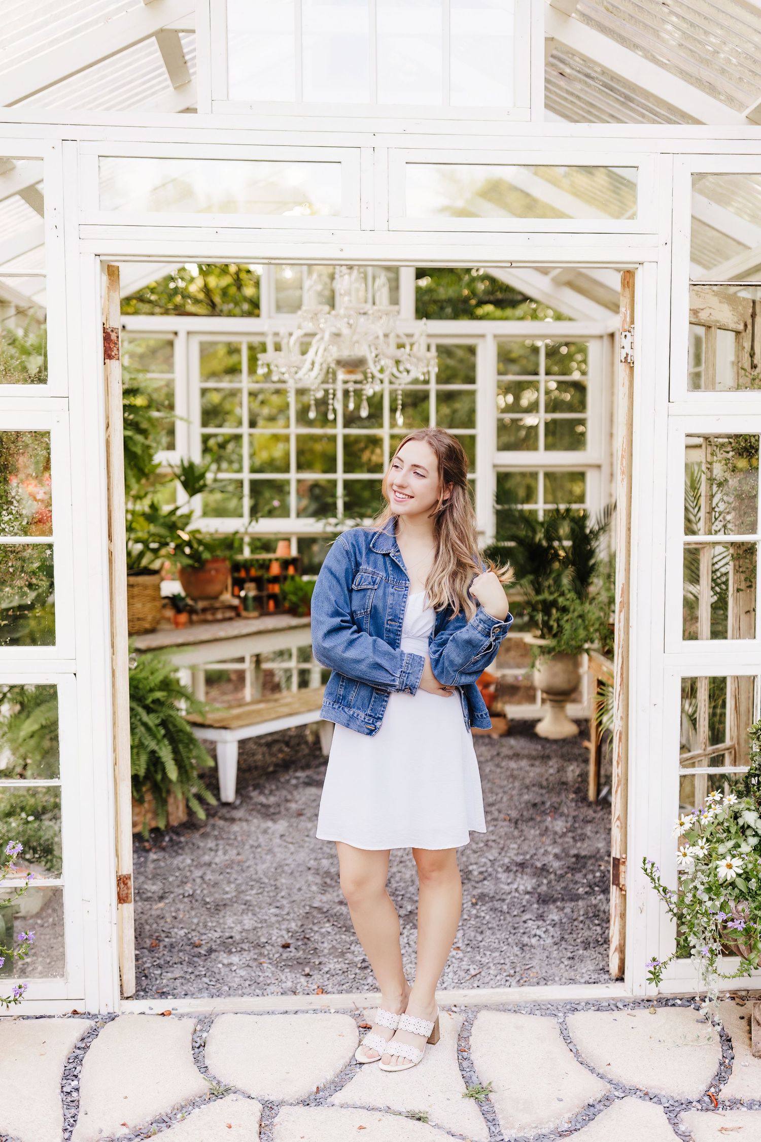Young woman in white dress and denim jacket smiling in doorway of charming glass greenhouse with chandelier.