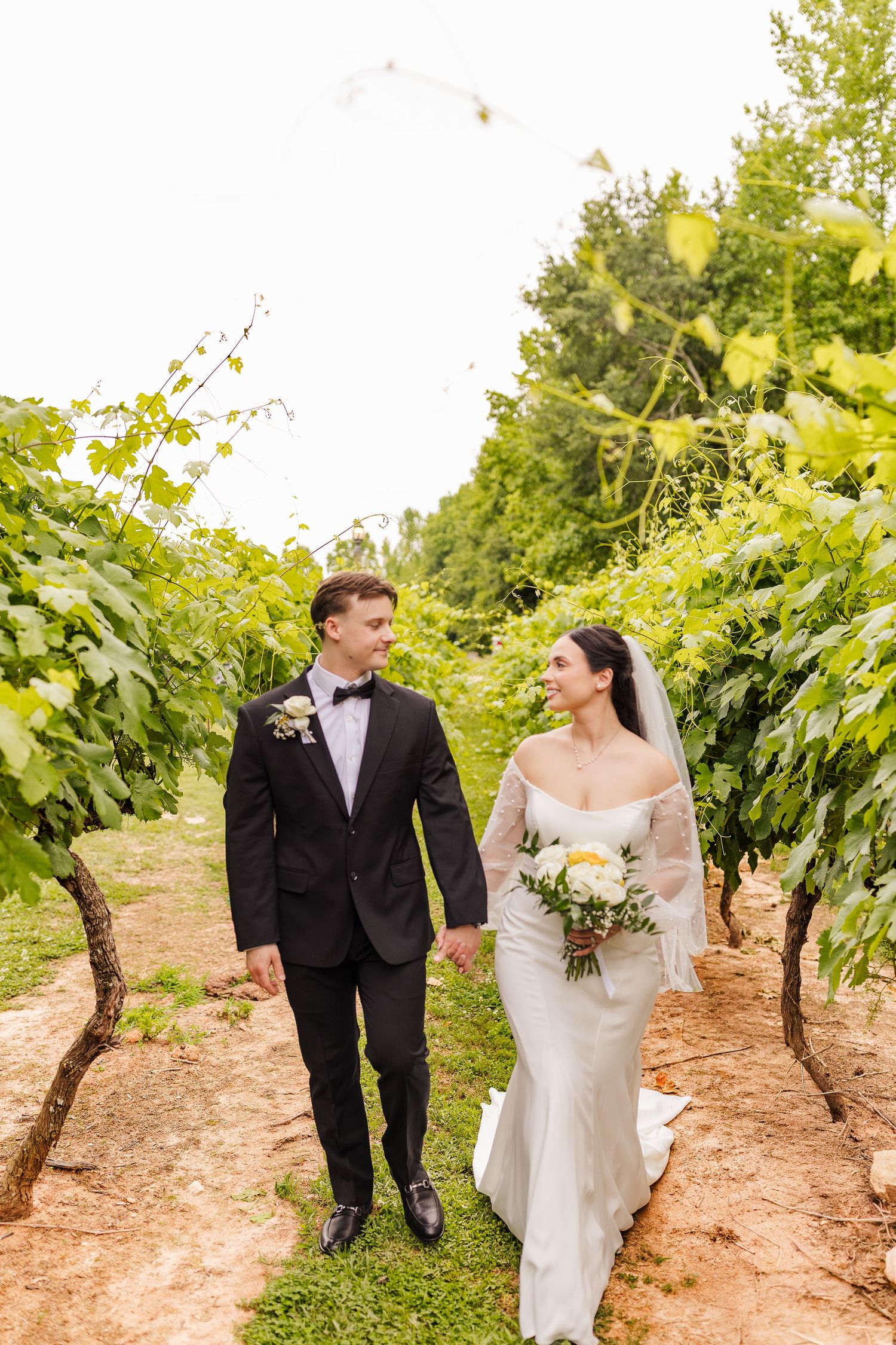 Bride and groom walk hand-in-hand through lush vineyard rows, bride holding white bouquet in elegant gown.