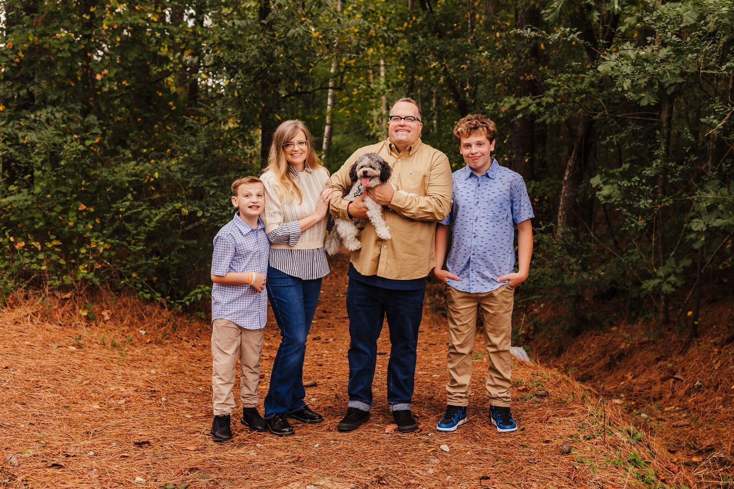 Family of four with dog posing for portrait on dirt trail in wooded setting.