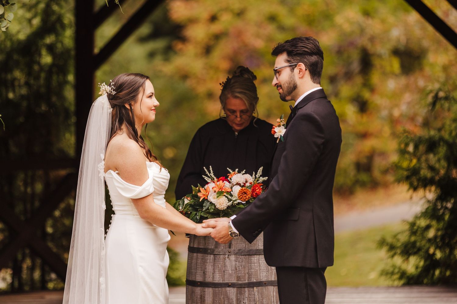 A wedding ceremony takes place outdoors with autumn foliage in the background and a rustic wine barrel altar decoration.
