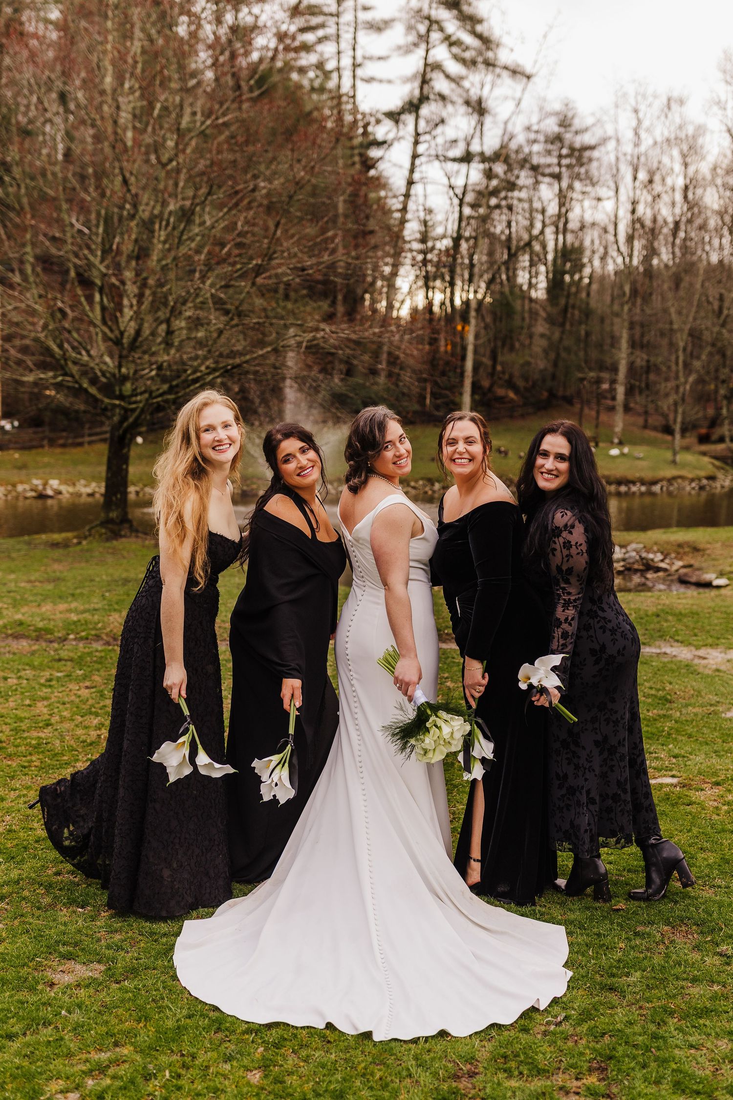 Bride in white gown poses with four bridesmaids in black dresses holding bouquets at outdoor fall wedding.