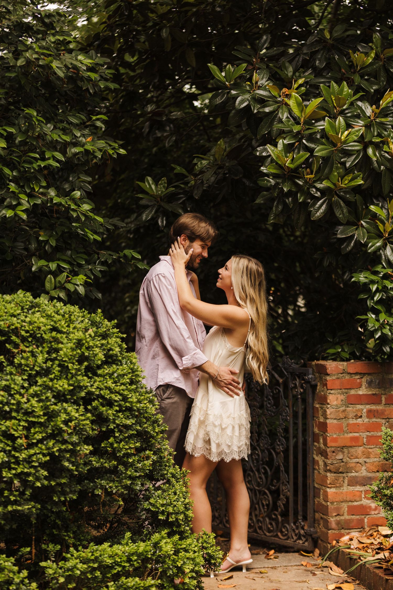 Couple embracing romantically in a lush garden near a brick wall and iron gate, woman in white lace dress, man in lavender shirt.