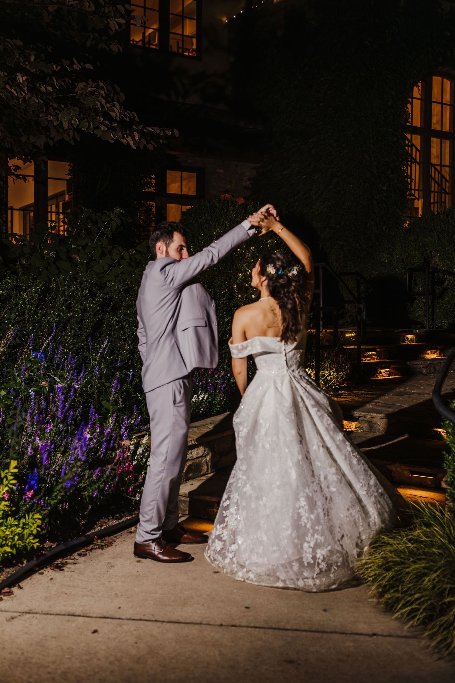 Bride and groom dance outdoors at night, groom twirling bride in lace gown under warm garden lights.