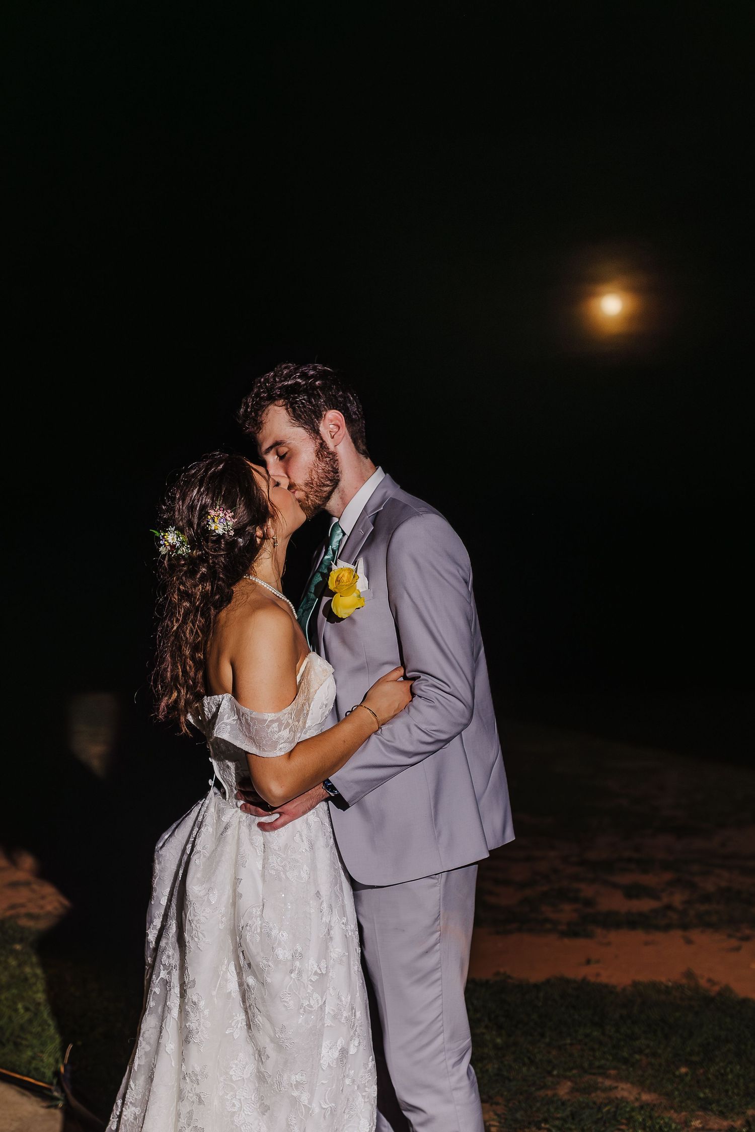 Bride and groom share a romantic kiss under a glowing full moon at their outdoor night wedding.
