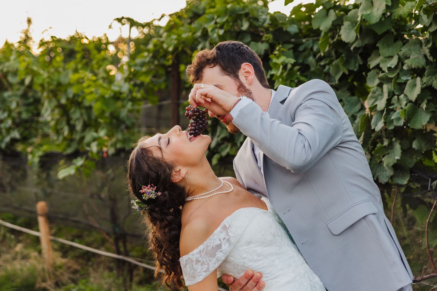 Bride and groom share grapes in a lush vineyard, bride in white off-shoulder gown, groom in light blue suit.