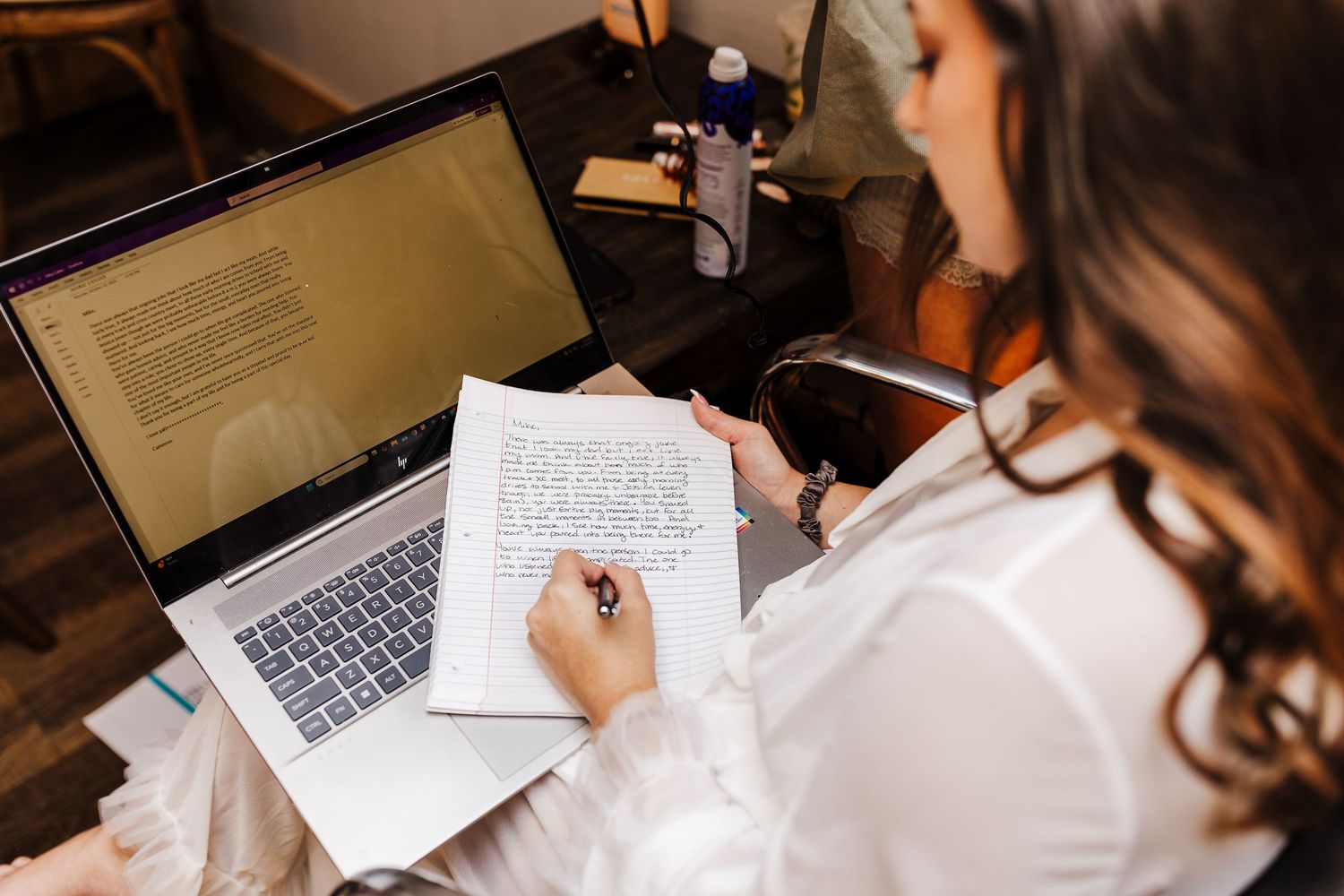 Student taking handwritten notes while studying at a desk with laptop and papers spread out.