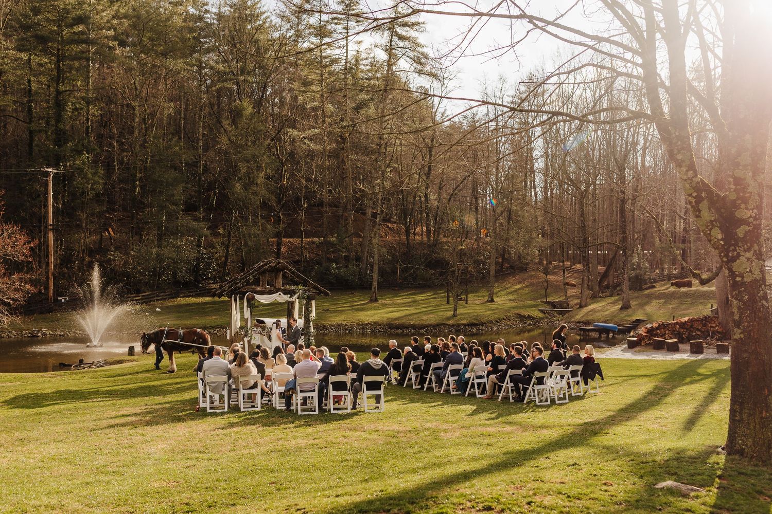 Outdoor wedding ceremony on lush green lawn surrounded by trees, with fountain pond and white chairs arranged for guests.
