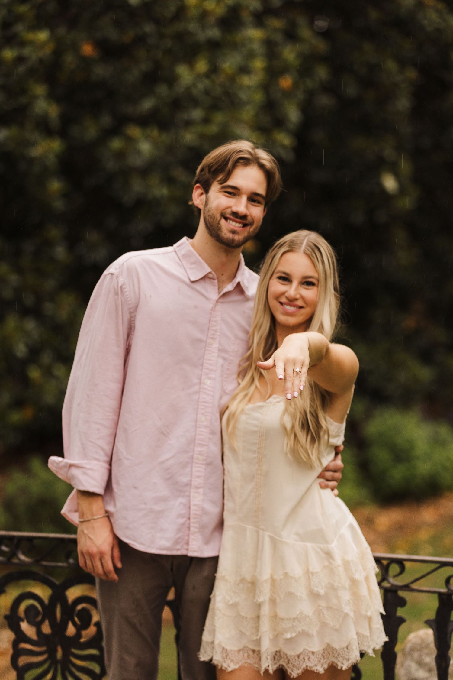 Happy couple posing outdoors, woman showing engagement ring in white lace dress beside man in pink shirt.