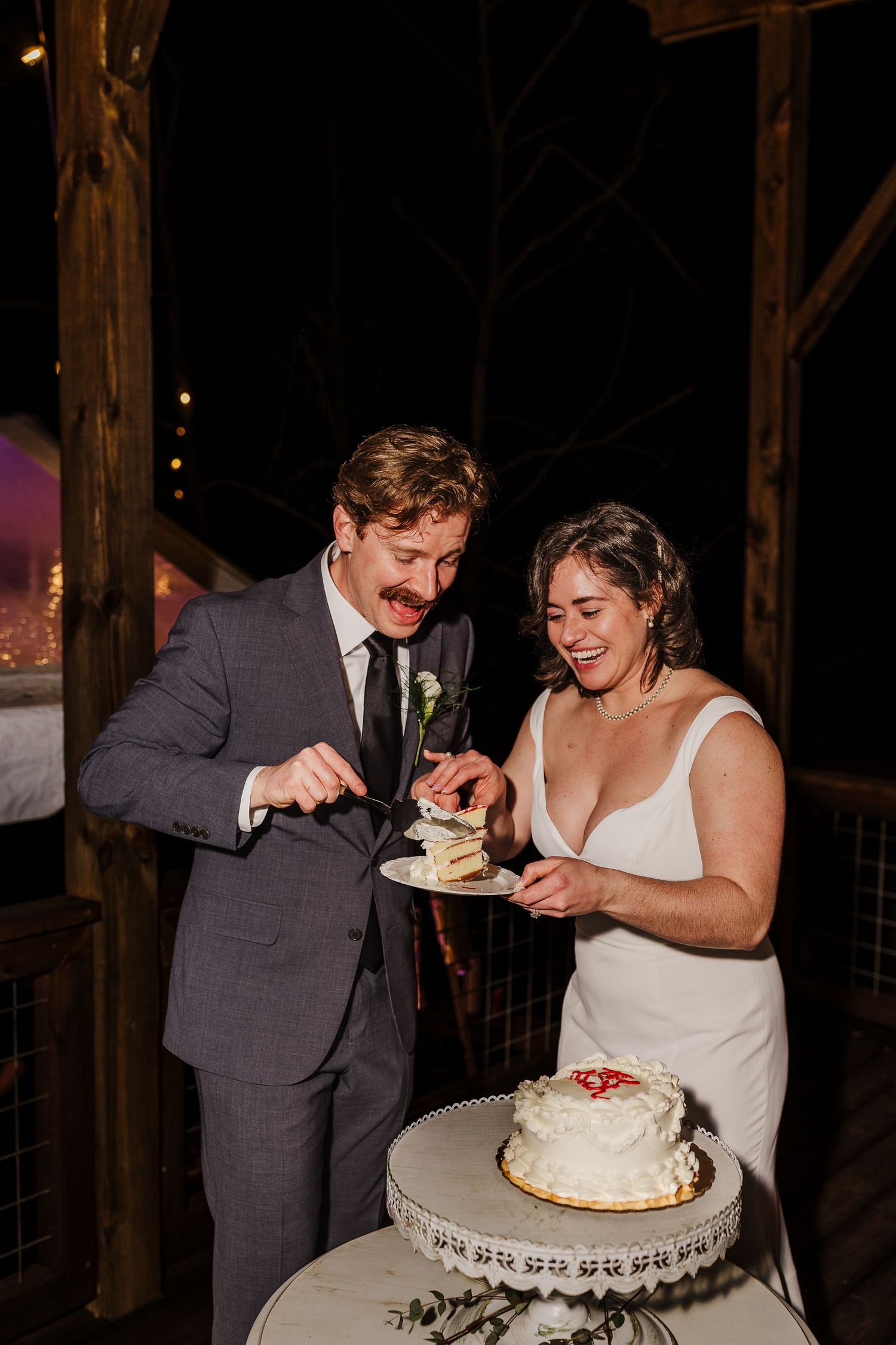 Bride and groom laughing while cutting white wedding cake together at outdoor evening reception.