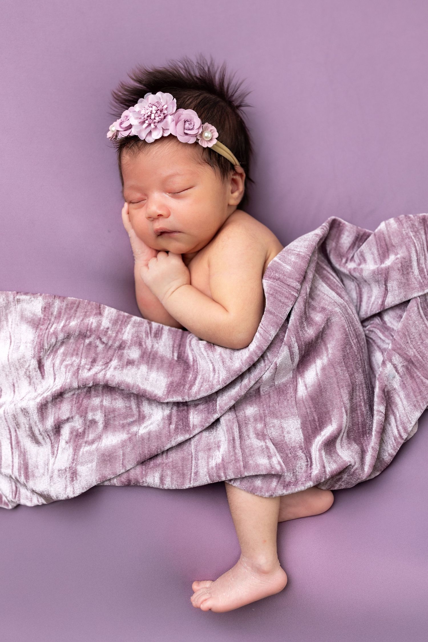 Sleeping newborn wrapped in pink silk fabric lying on purple backdrop with delicate floral headband.