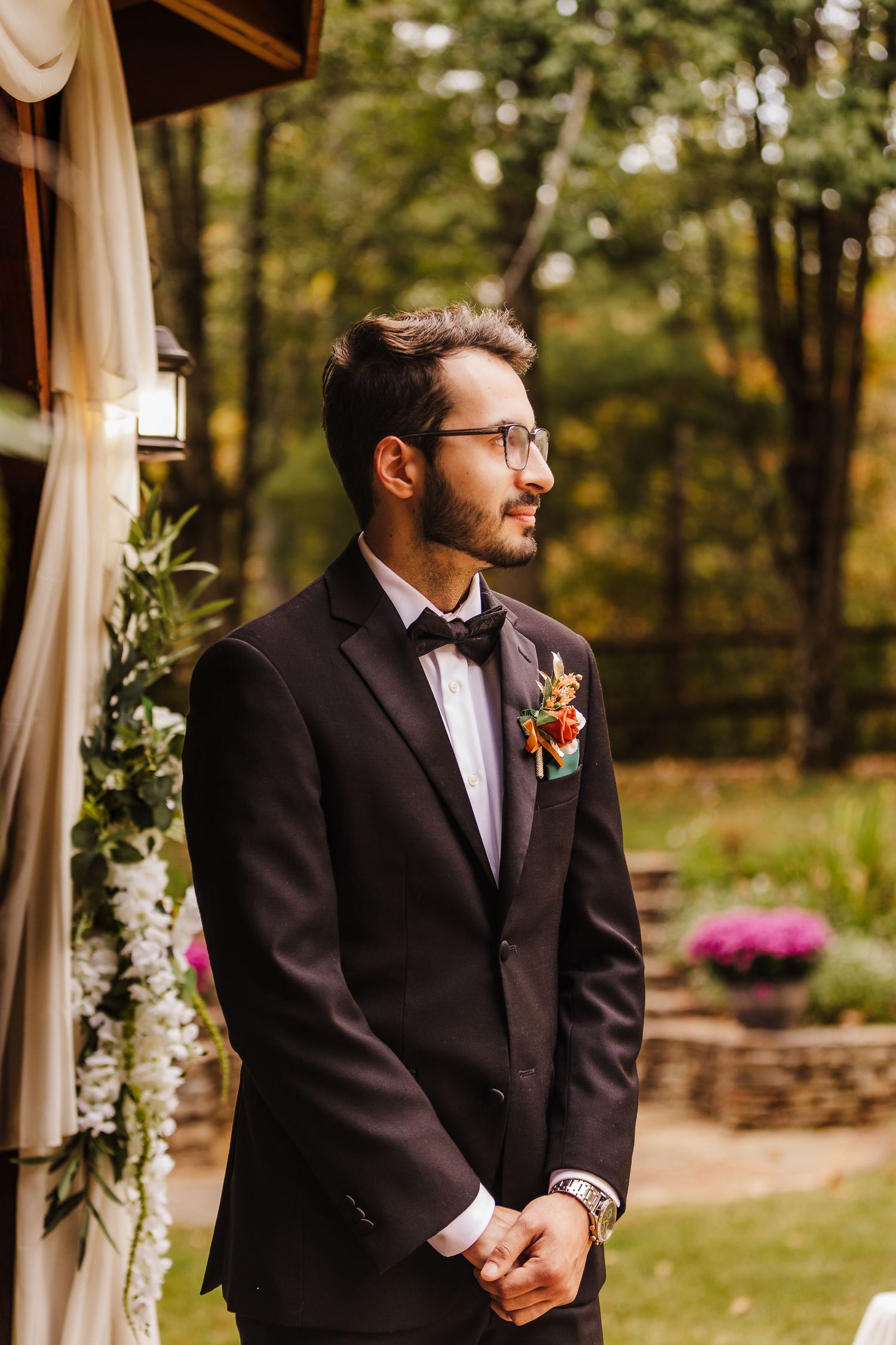 Groom in a classic black tuxedo waits expectantly at an outdoor wedding ceremony with draped fabric decor.