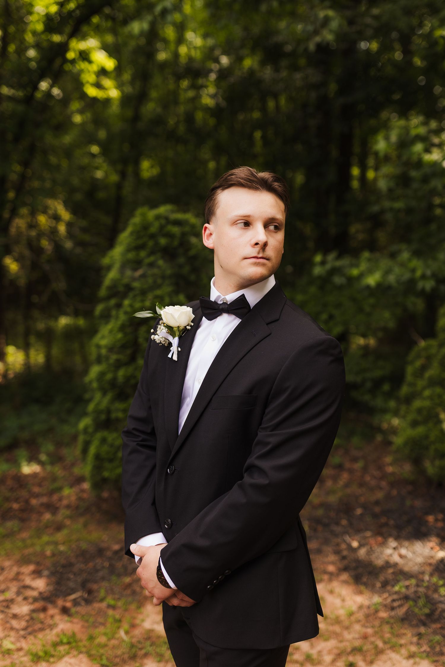 Elegant groom in black tuxedo with bow tie and white boutonniere posing outdoors in lush green forest setting.