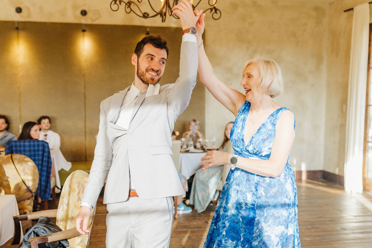 Groom in white suit dancing with elderly woman in blue dress at elegant wedding reception venue.