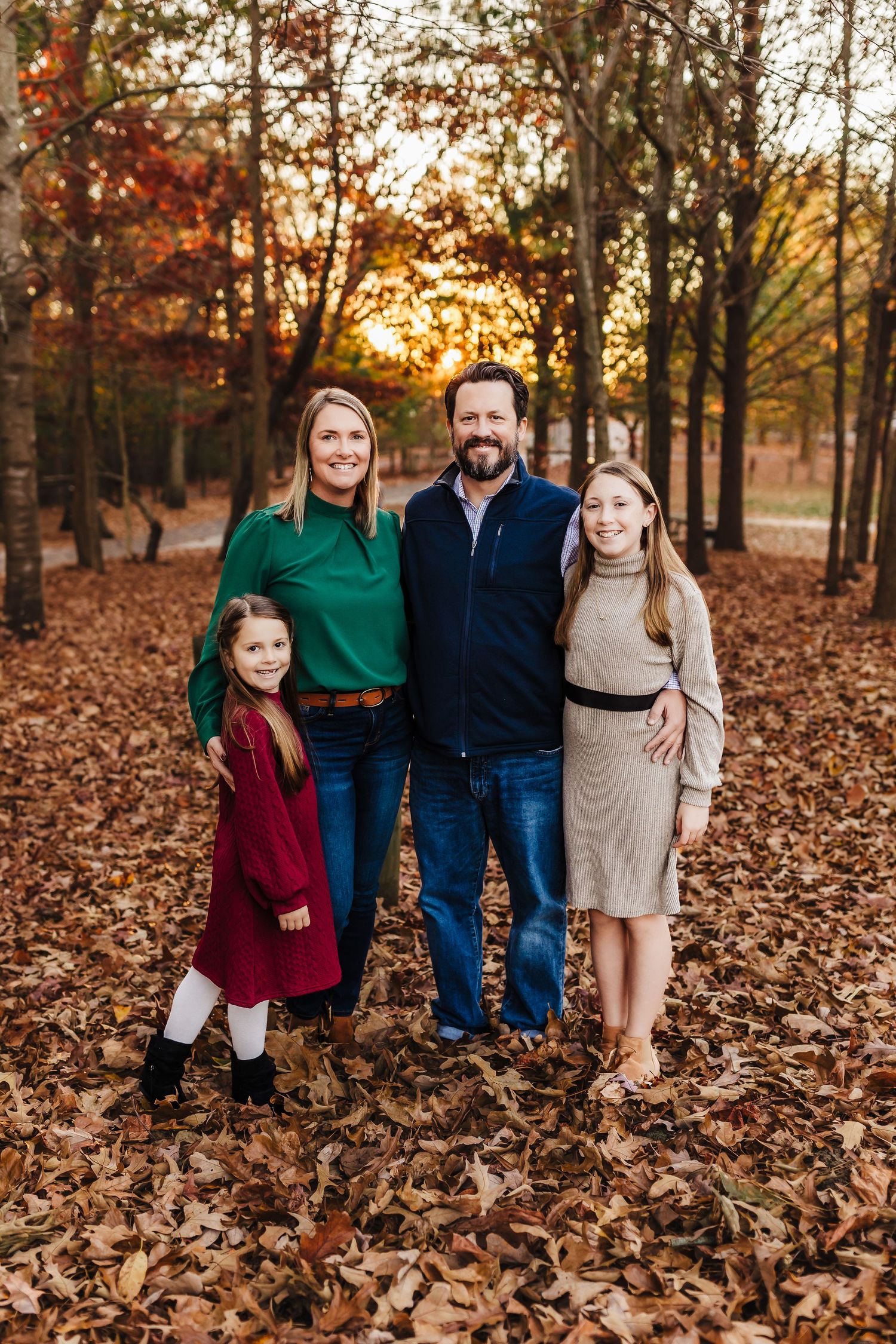 Family of four in coordinated fall outfits standing among fallen autumn leaves in a wooded setting.