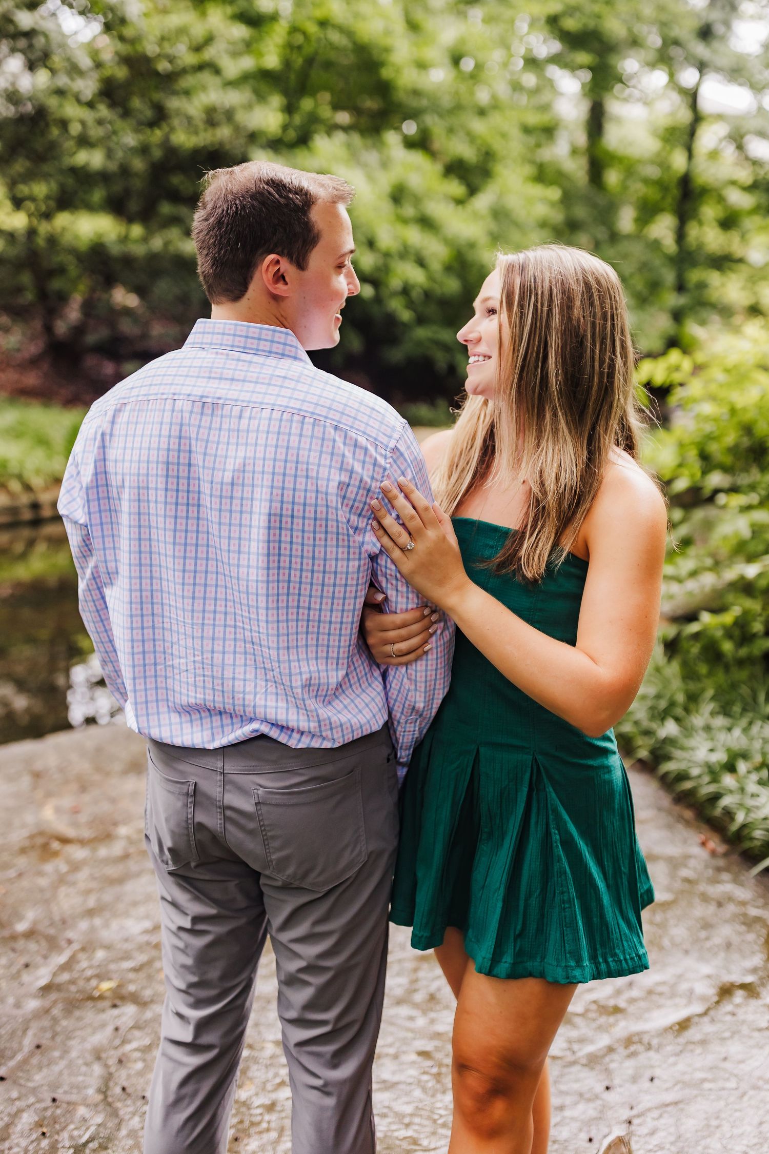 Couple sharing a romantic moment on a wooded path with woman in green dress embracing man in plaid shirt from behind.