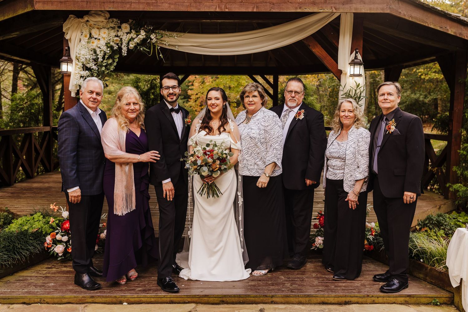 Wedding celebration group poses under draped canopy with floral arrangements at outdoor ceremony venue.