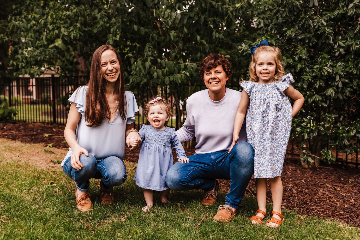A candid outdoor family portrait with adults and young children sitting together on mulch surrounded by greenery.