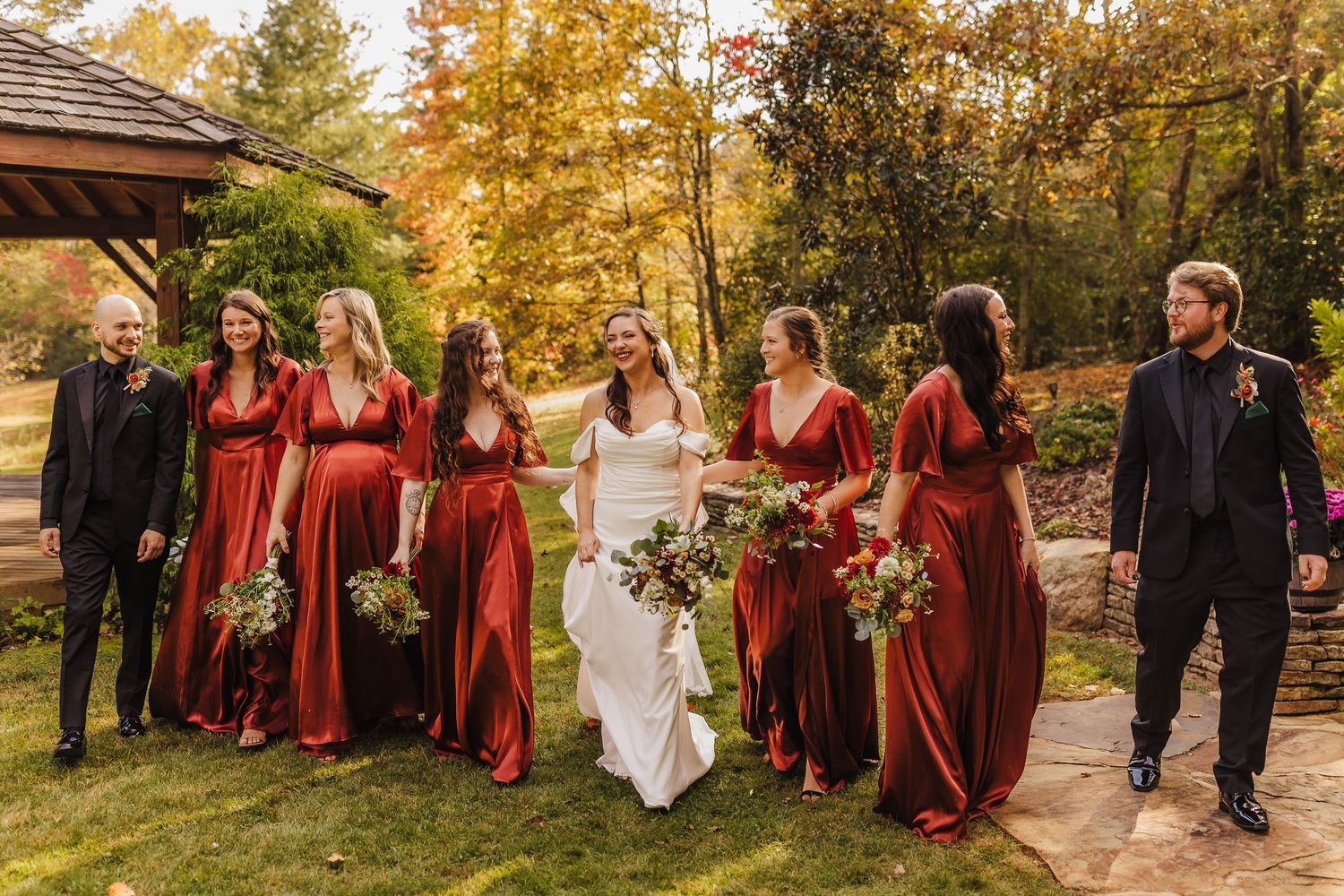 Wedding party in burgundy dresses and formal attire walking through autumn trees with golden foliage.