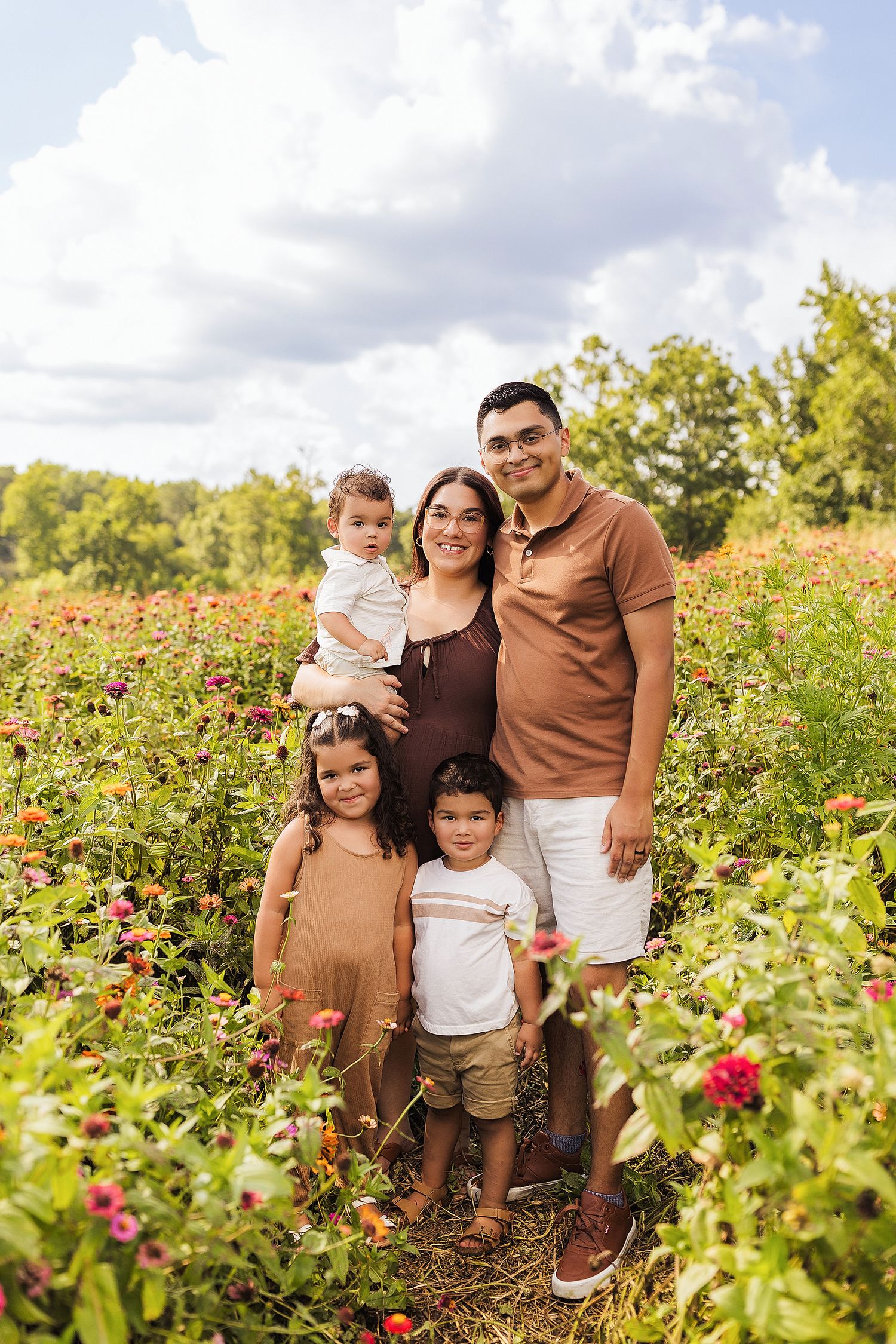 Family of five posing in a colorful flower field on a sunny day, with parents holding young children among blooming zinnias.