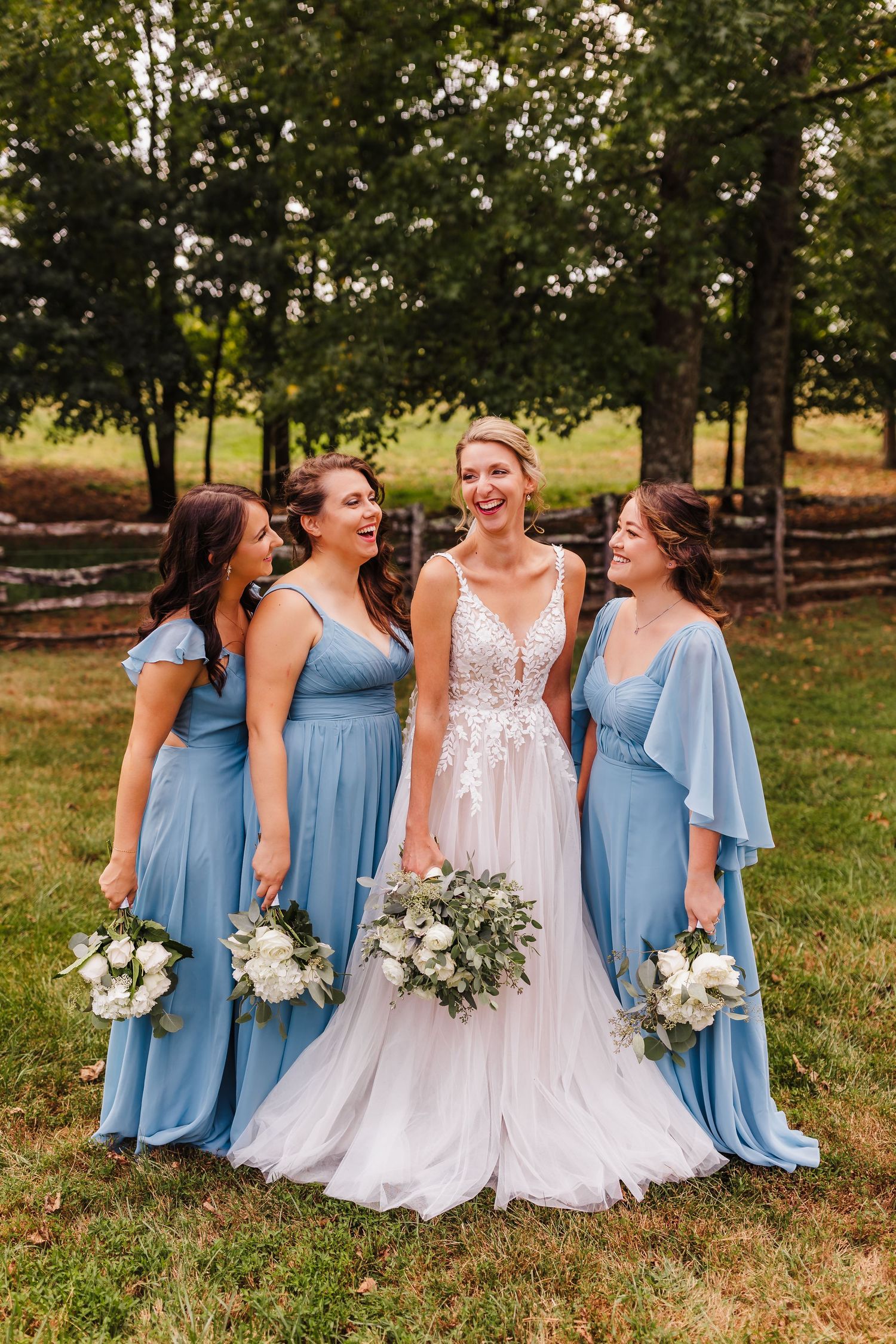 Wedding party in light blue bridesmaid dresses posing outdoors with rustic fence backdrop.
