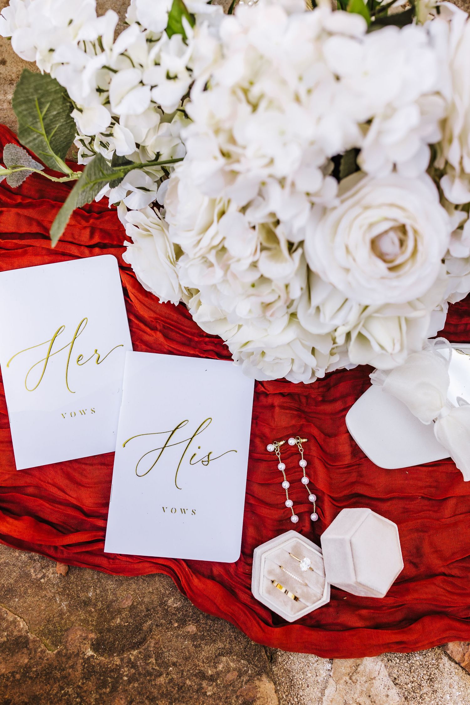 Wedding stationery featuring 'Her' cards and white roses arranged on red fabric with decorative accessories.