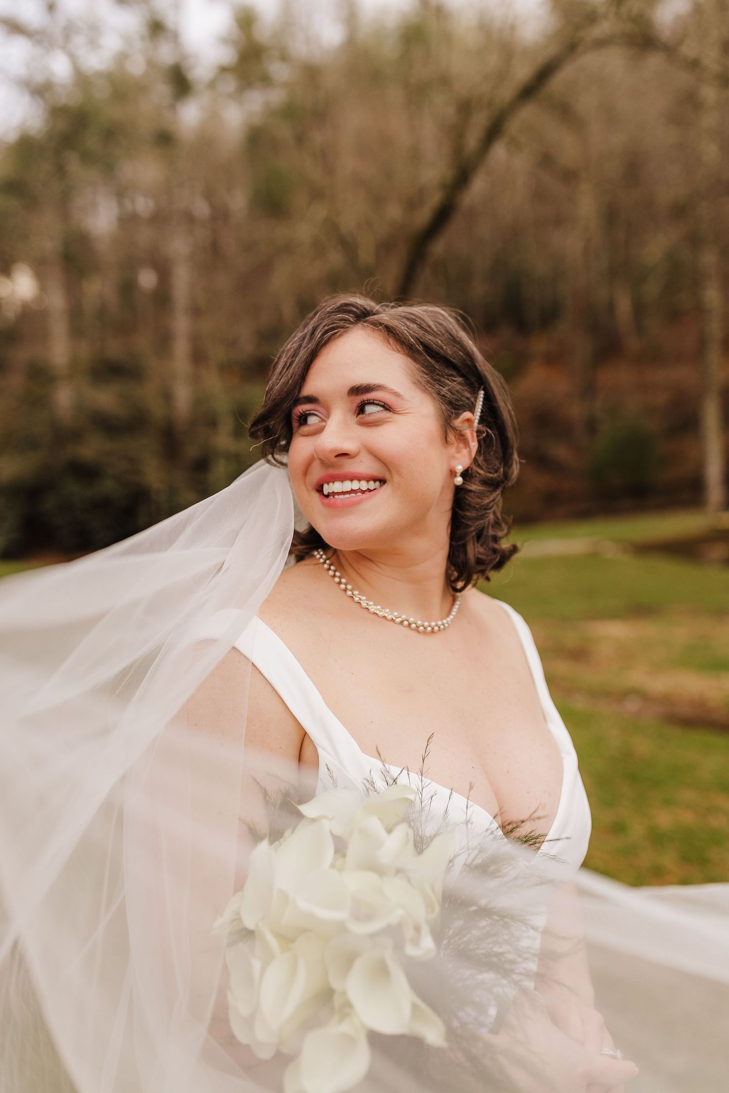 Smiling bride in white gown and pearl necklace holds bouquet as veil flows in breeze outdoors.