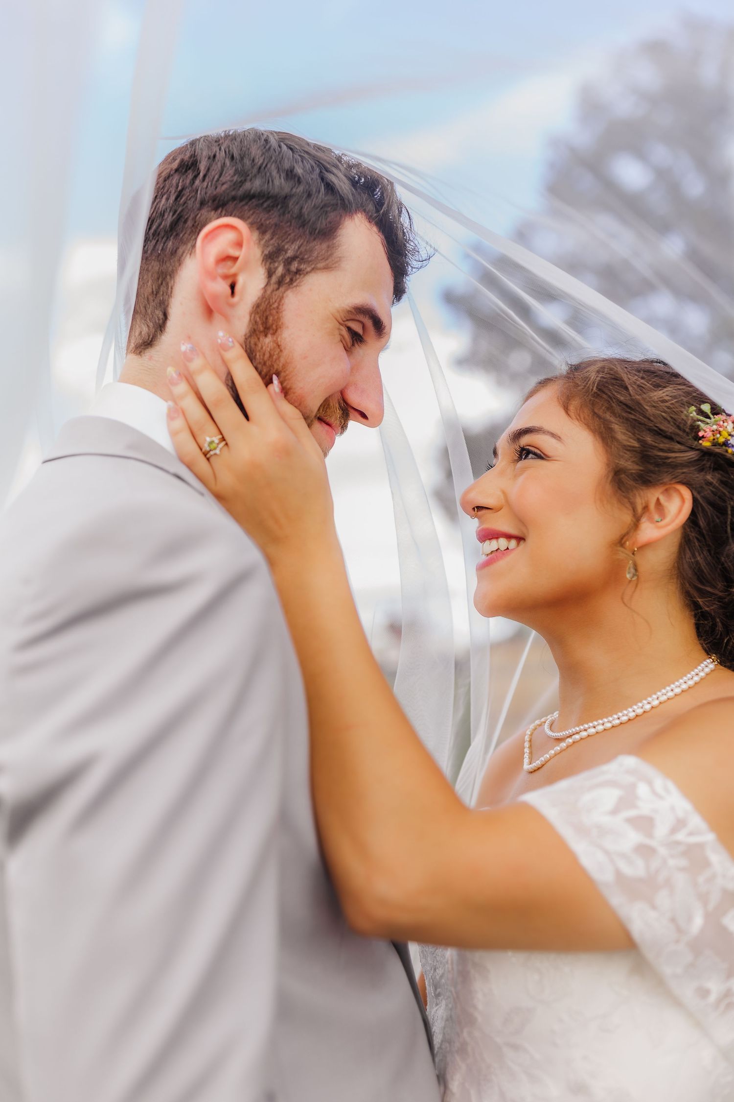 A smiling bride in a white lace dress and pearl necklace gazes at her groom under a flowing veil.