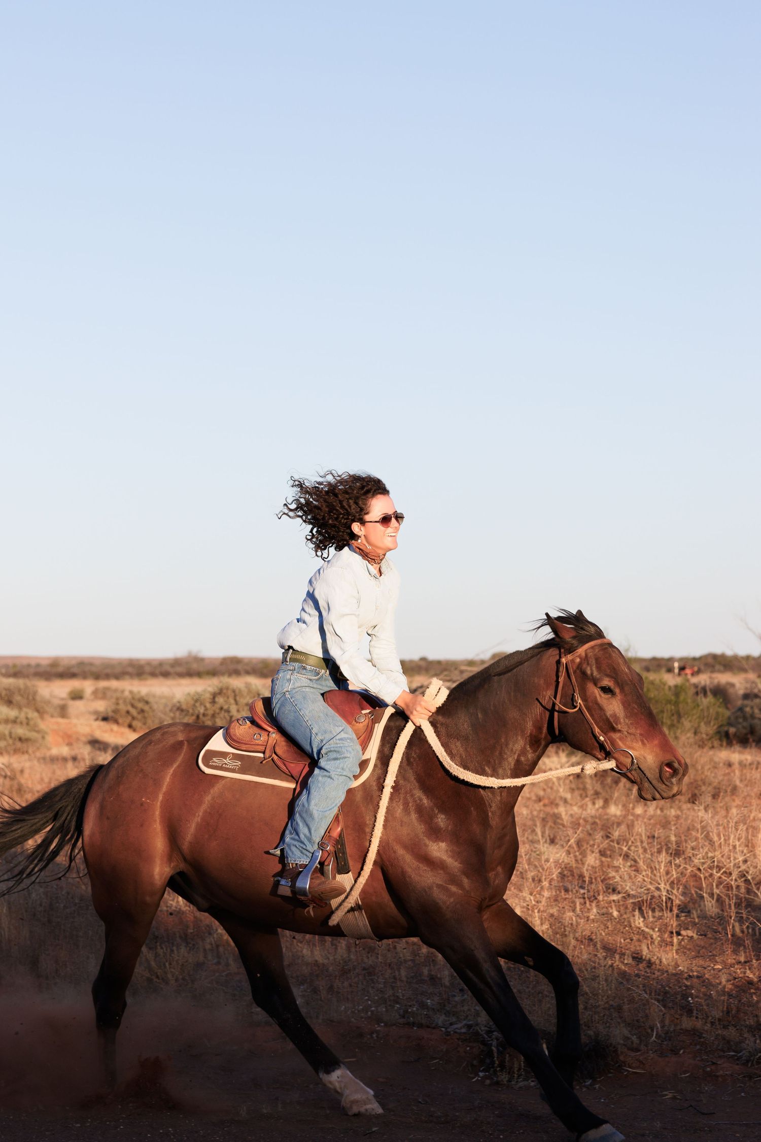 A rider on a brown horse gallops across a dusty open field at sunset in the desert.