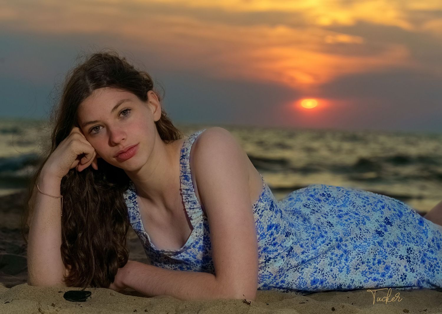 Young woman in blue floral dress lying on sandy beach gazing at camera with vibrant sunset behind her.