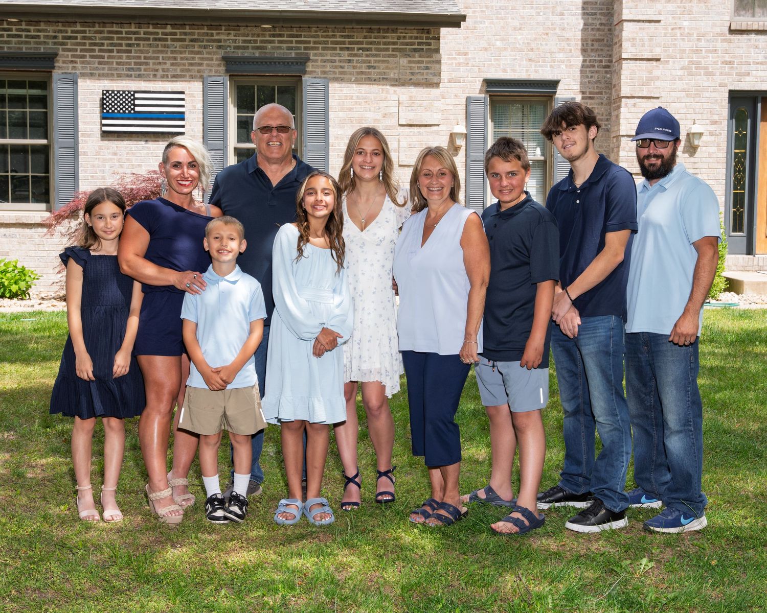 A large extended family smiles together outdoors on a green lawn in front of a brick home.