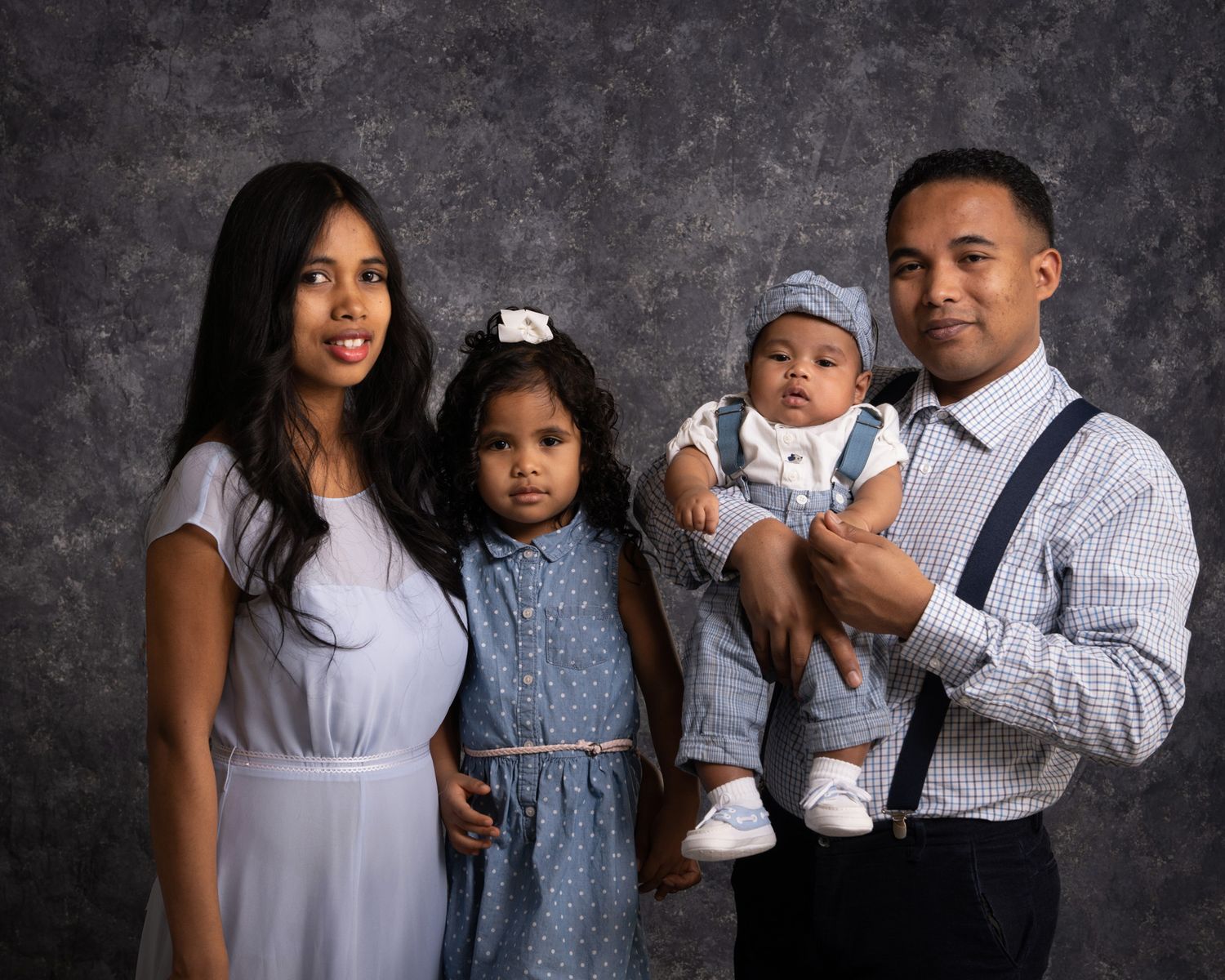 Family of four with toddler girl and baby boy in coordinating blue outfits studio portrait.