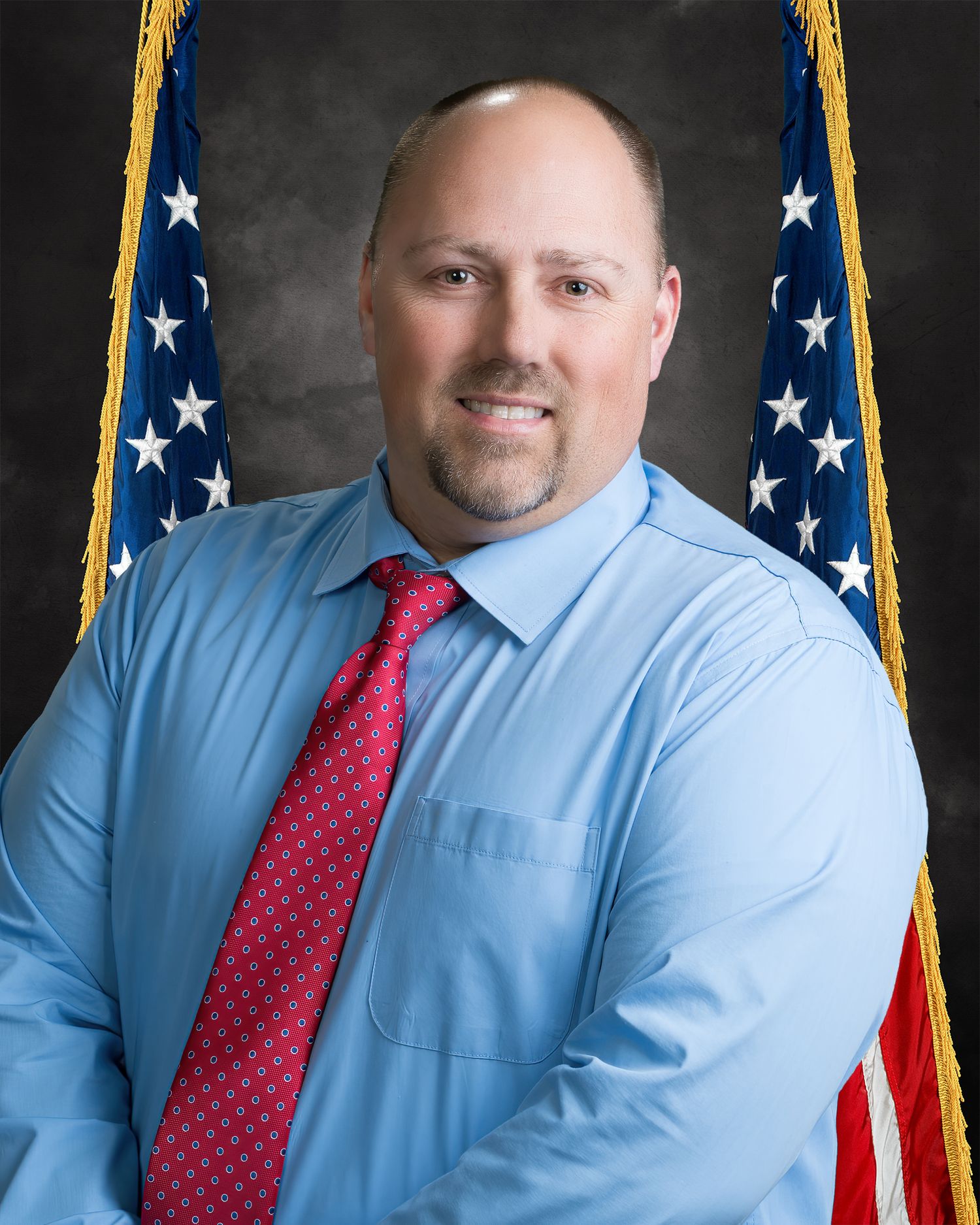 Official portrait of a man in blue shirt and red tie smiling before an American flag background.