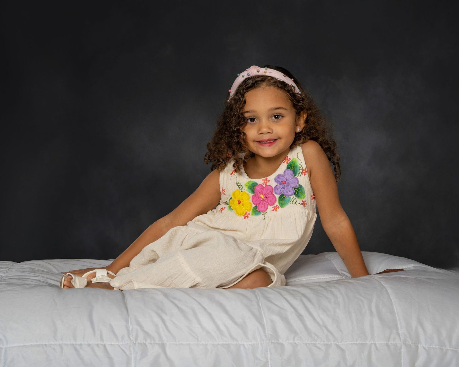 Adorable young girl in floral white dress sitting on white surface smiling.