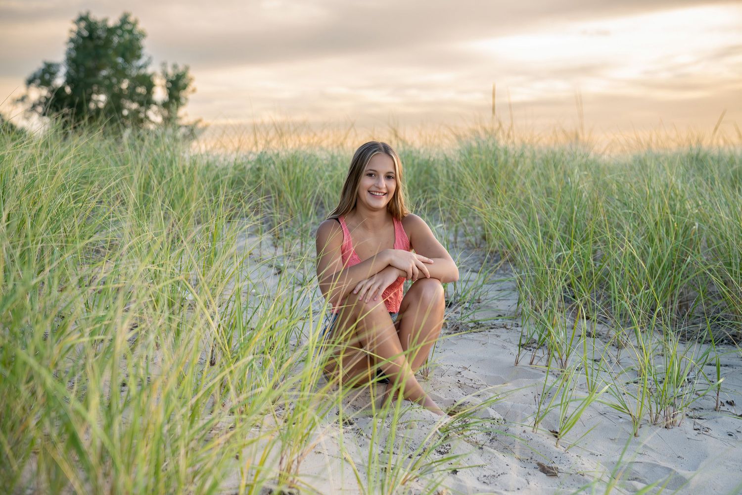 Smiling teen girl in coral top sitting among tall beach grass dunes at golden hour.