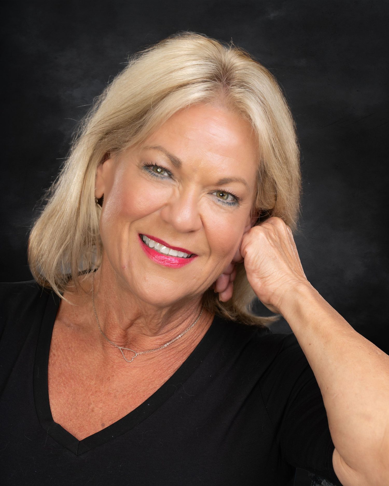 Blonde woman in black V-neck top posing with hand near face on dark background.
