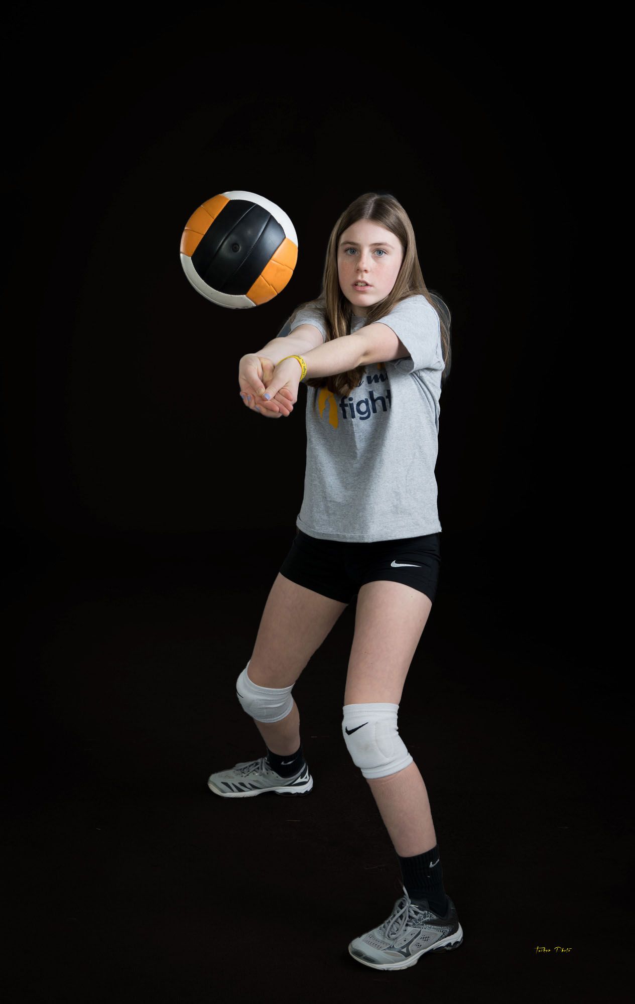 Young female volleyball player bumping a ball wearing a gray fight shirt, Nike shorts, and knee pads.