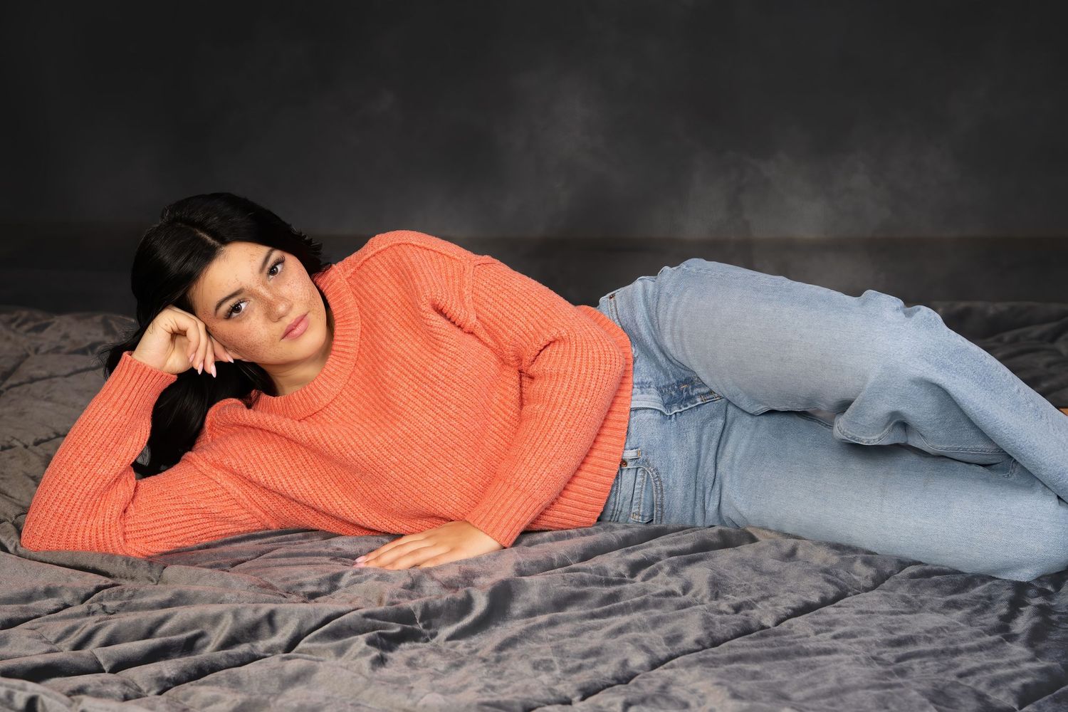 Woman in coral sweater and jeans posing on gray bedding surface.