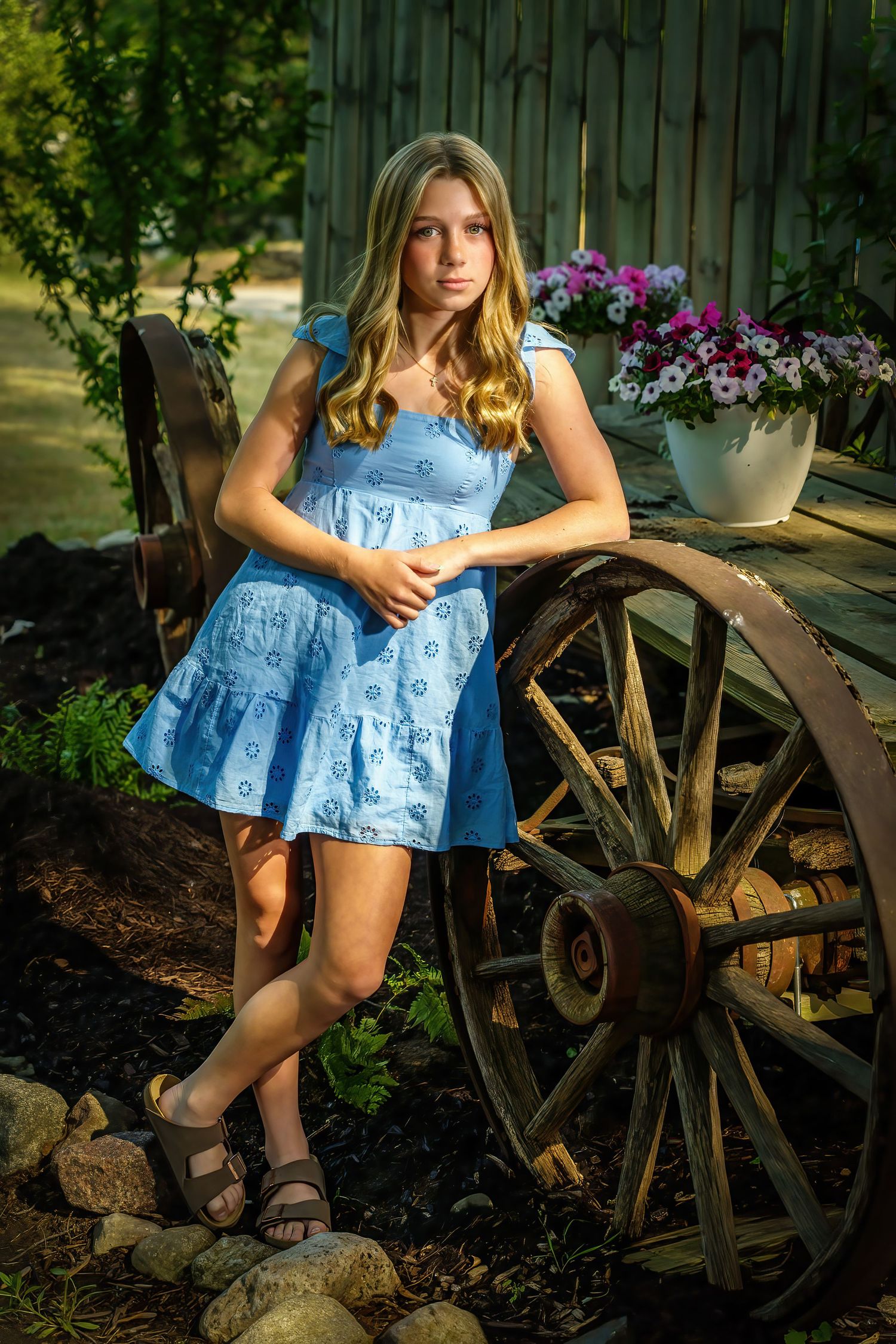 Teen girl in a light blue sundress leaning against a rustic wooden wagon wheel in an outdoor garden setting.