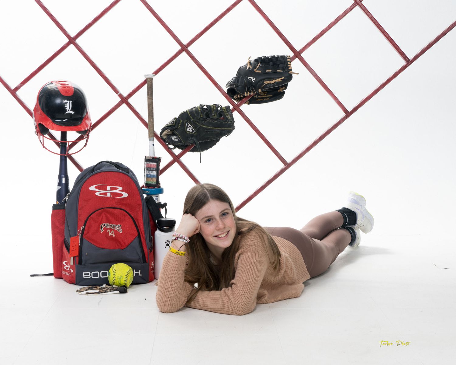 Female softball player poses with her Pirates gear, backpack, helmet, gloves, and bat on a white studio background.